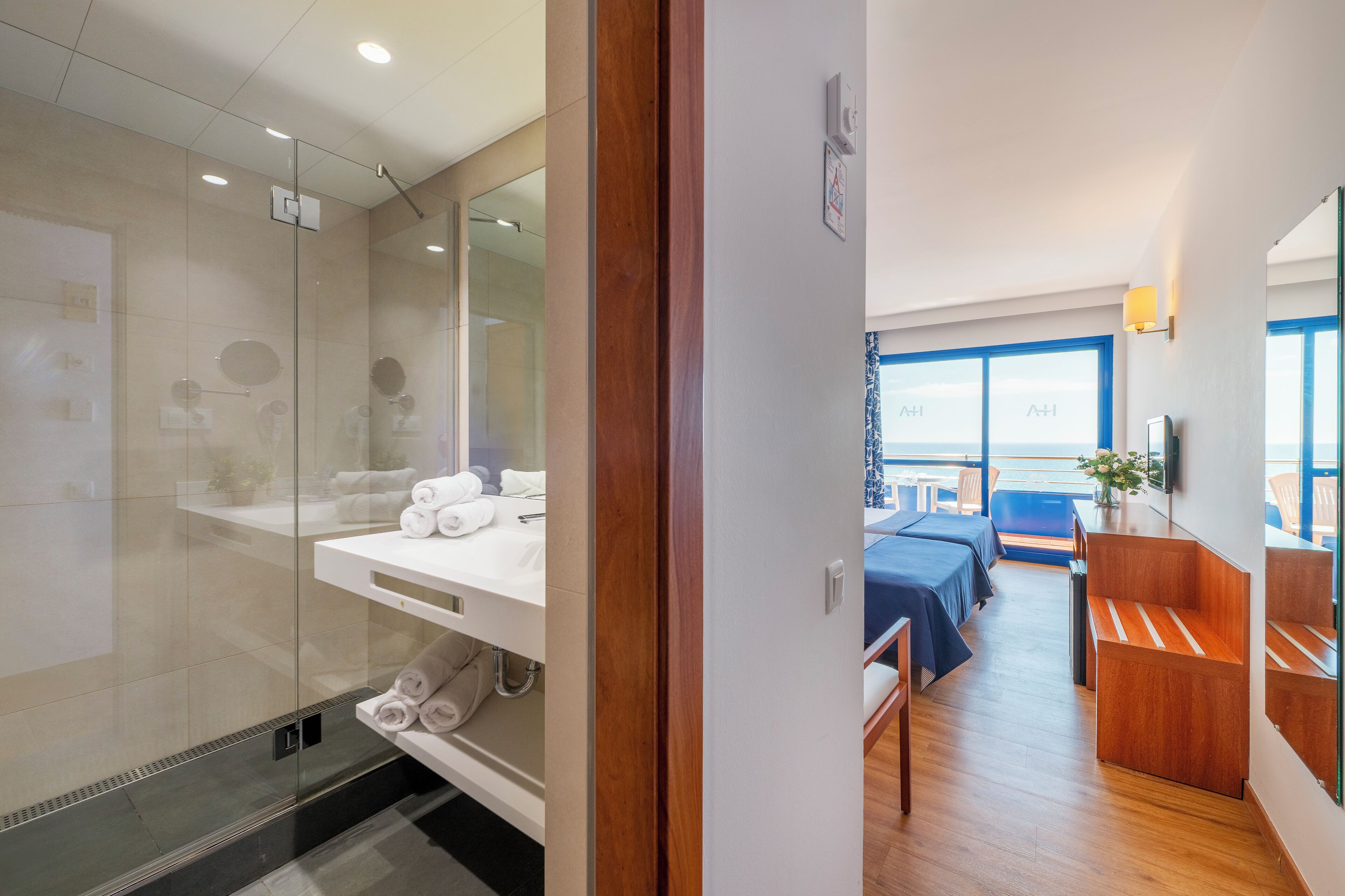 Een moderne hotelkamer met een glazen douchecabine, een houten wastafelmeubel, een bed met een blauwe spreideken en een balkon met uitzicht.