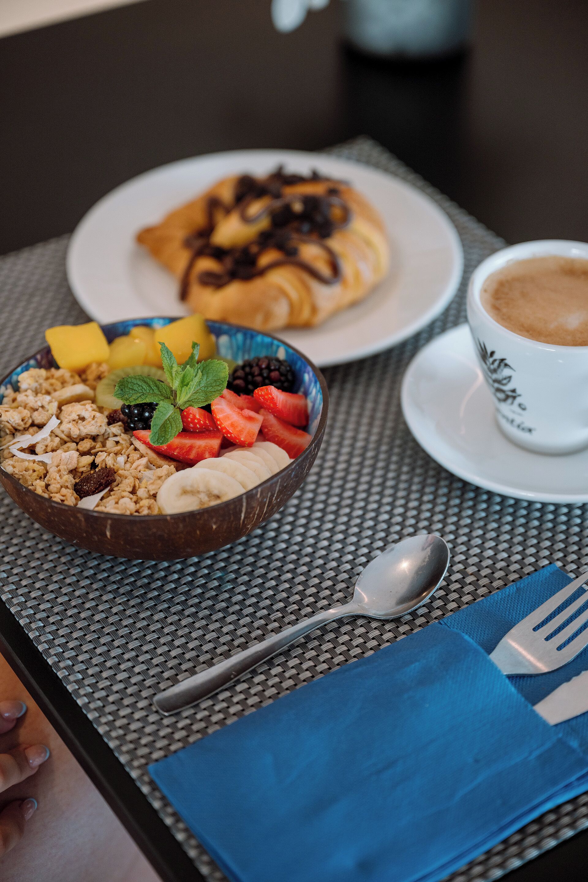 A breakfast setting with a bowl of cereal, fruits, a plate of pancakes, and a cup of coffee on a table.