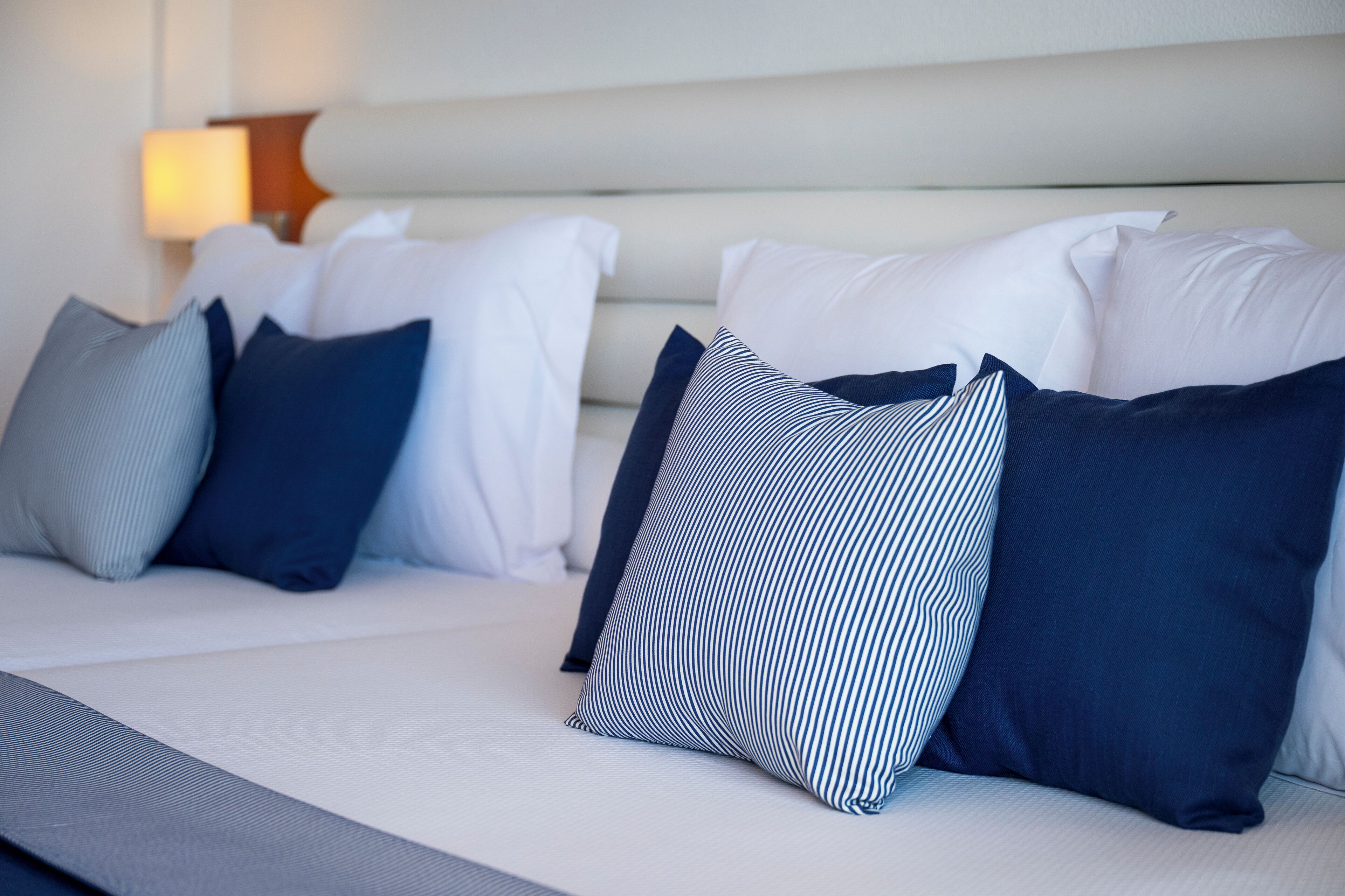 Een net opgemaakt bed met witte en marineblauwe kussens, een wit hoofdbord en een bedlampje.