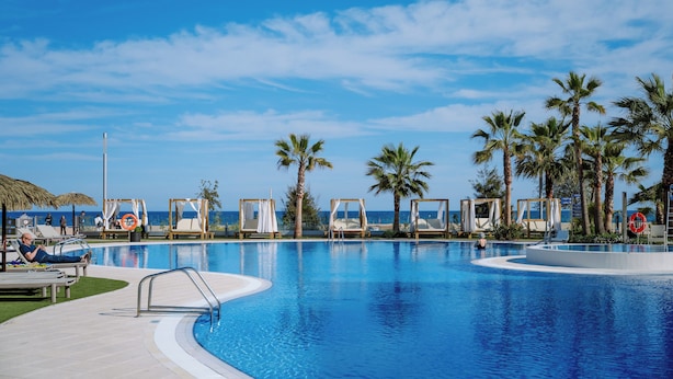 Ein Poolbereich mit Liegestühlen und Palmen, mit Blick auf einen Strand.