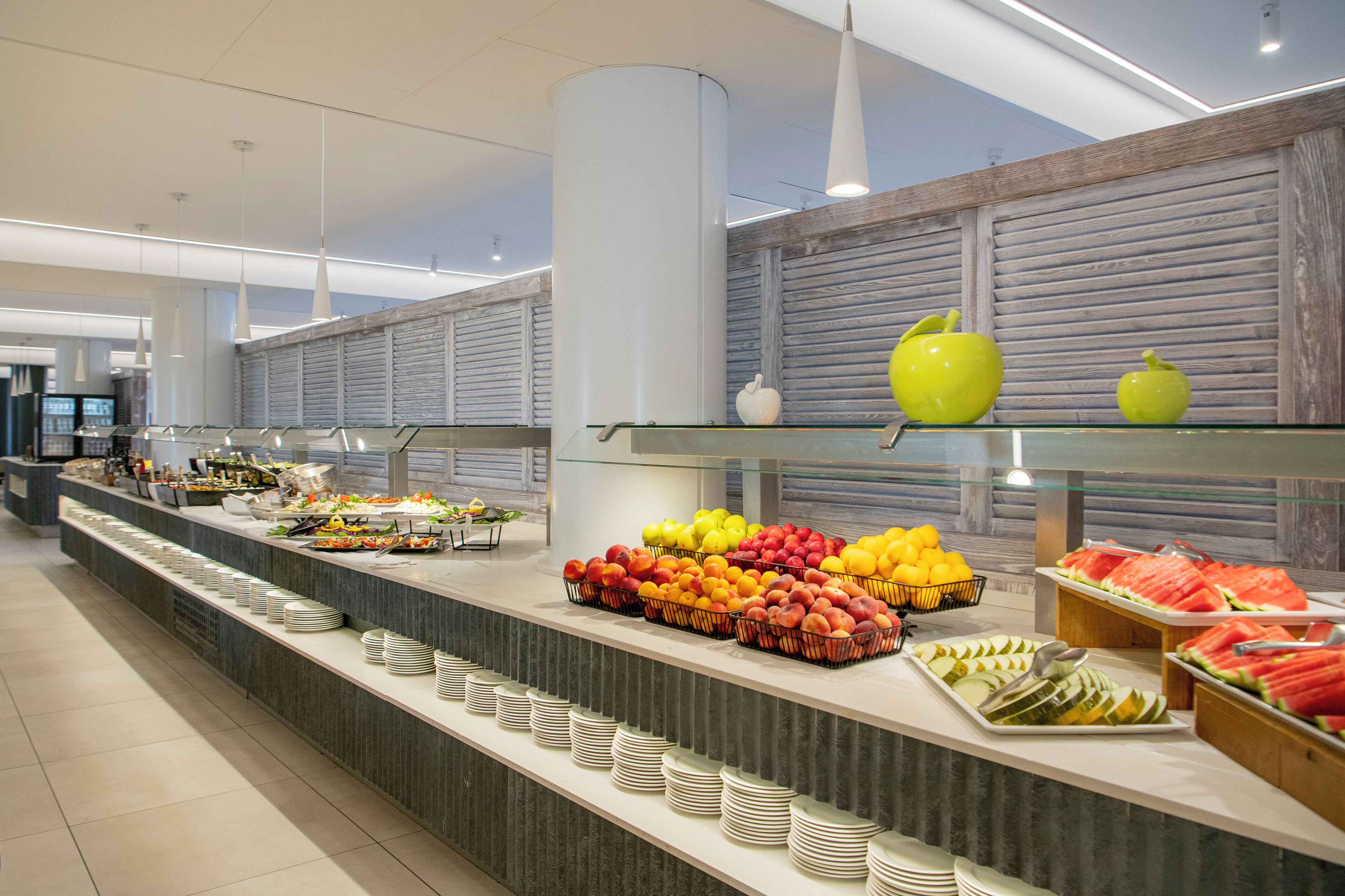 Ein Buffet mit verschiedenen Früchten und Gemüsesorten, darunter Äpfel, Orangen und Wassermelone, präsentiert auf einer Theke mit Glastrensen.