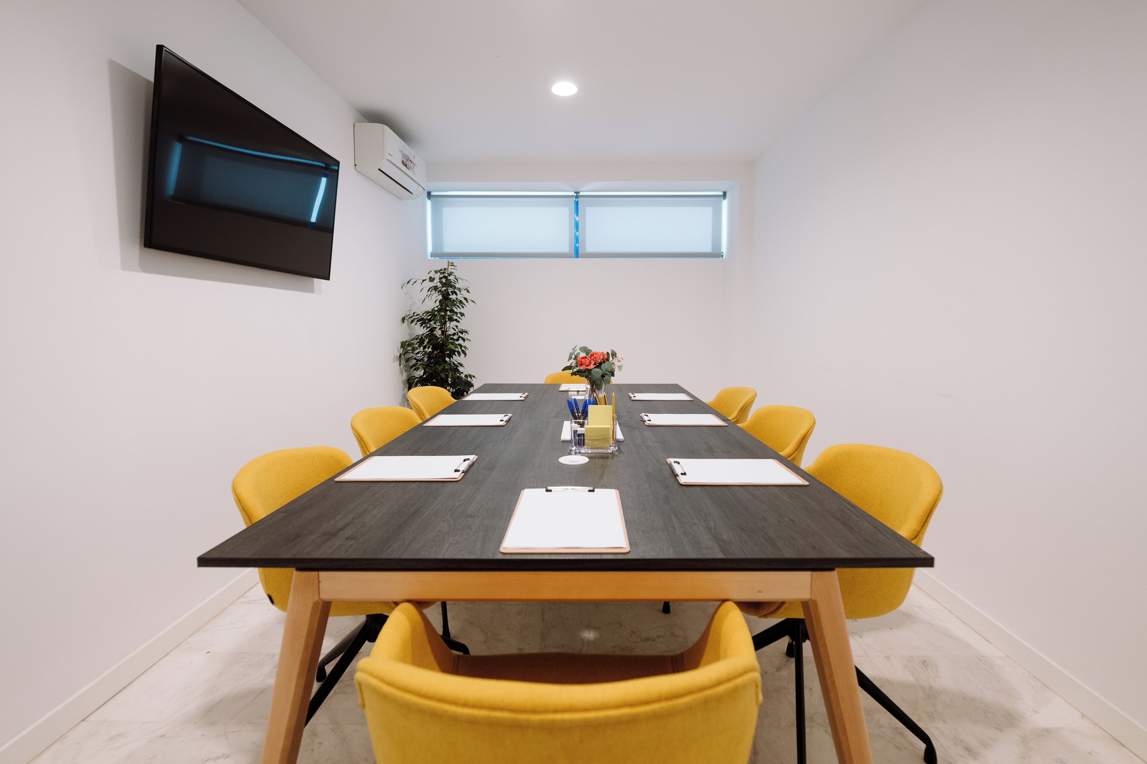 Un salón de conferencias con una larga mesa, sillas amarillas, un televisor montado y una planta en maceta.