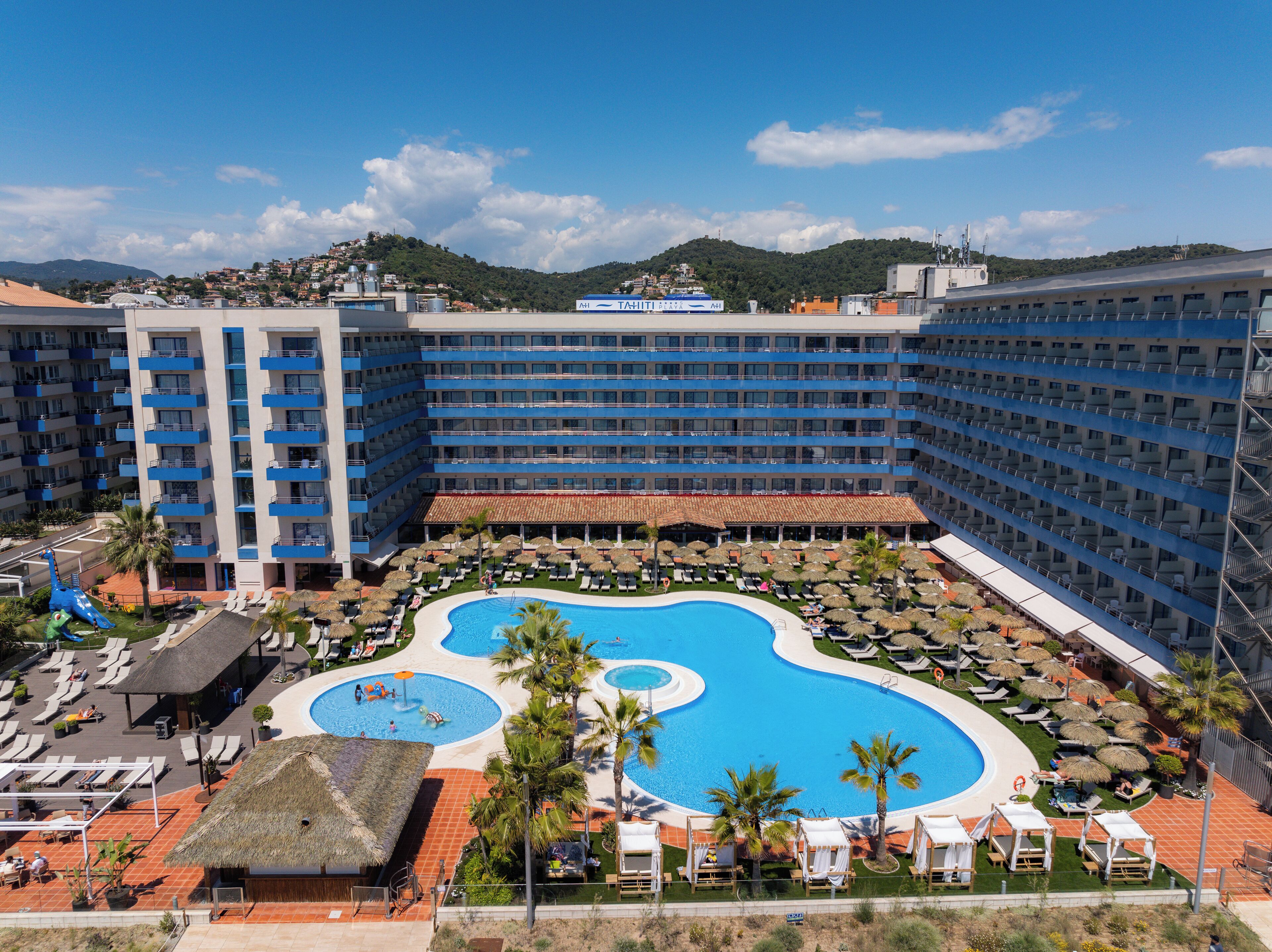 A hotel with a large pool area, multiple balconies, and a view of the surrounding hills.