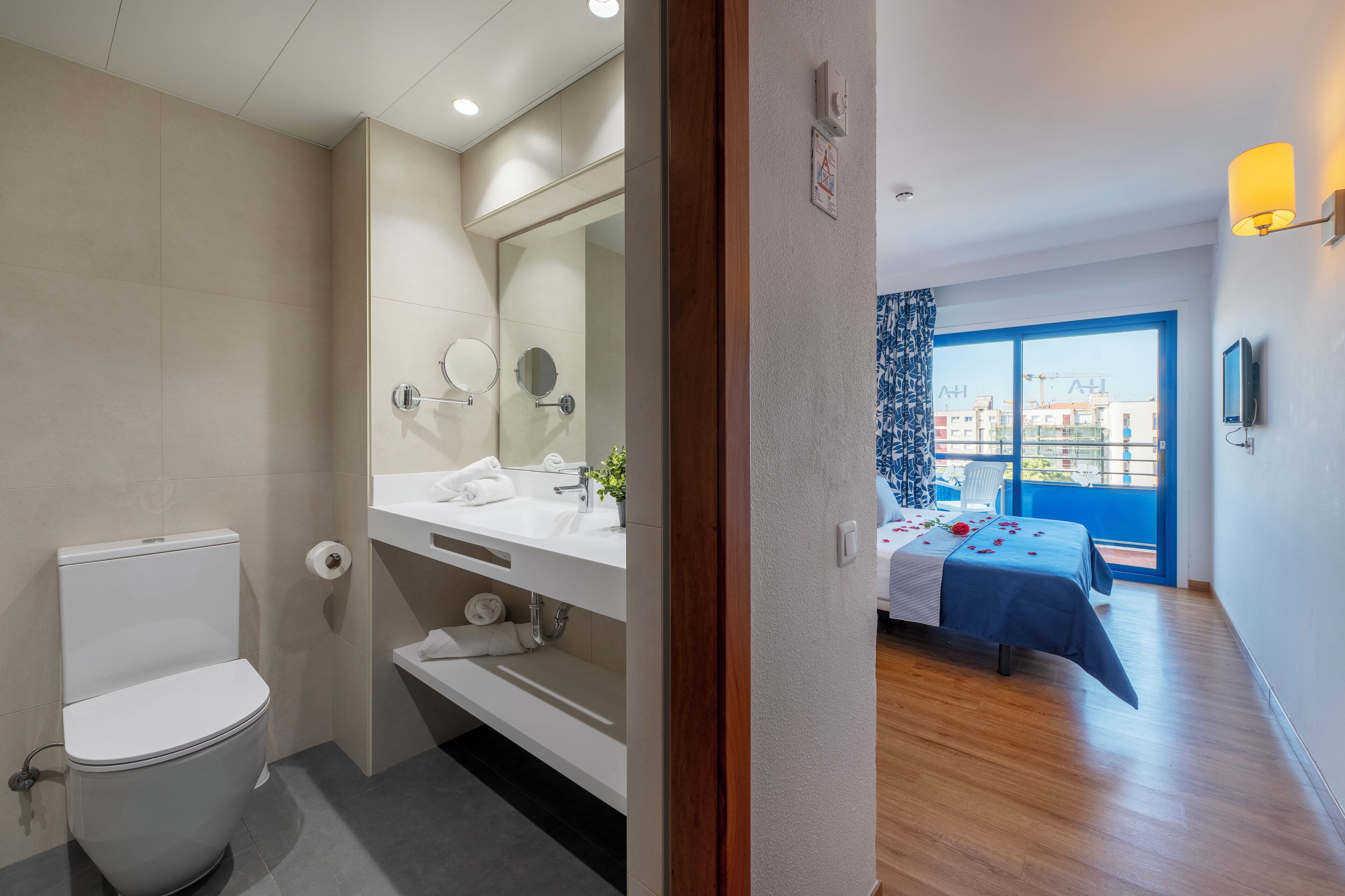 A modern bathroom with a toilet, sink, and mirror. There is a balcony with a bed and a view of buildings.