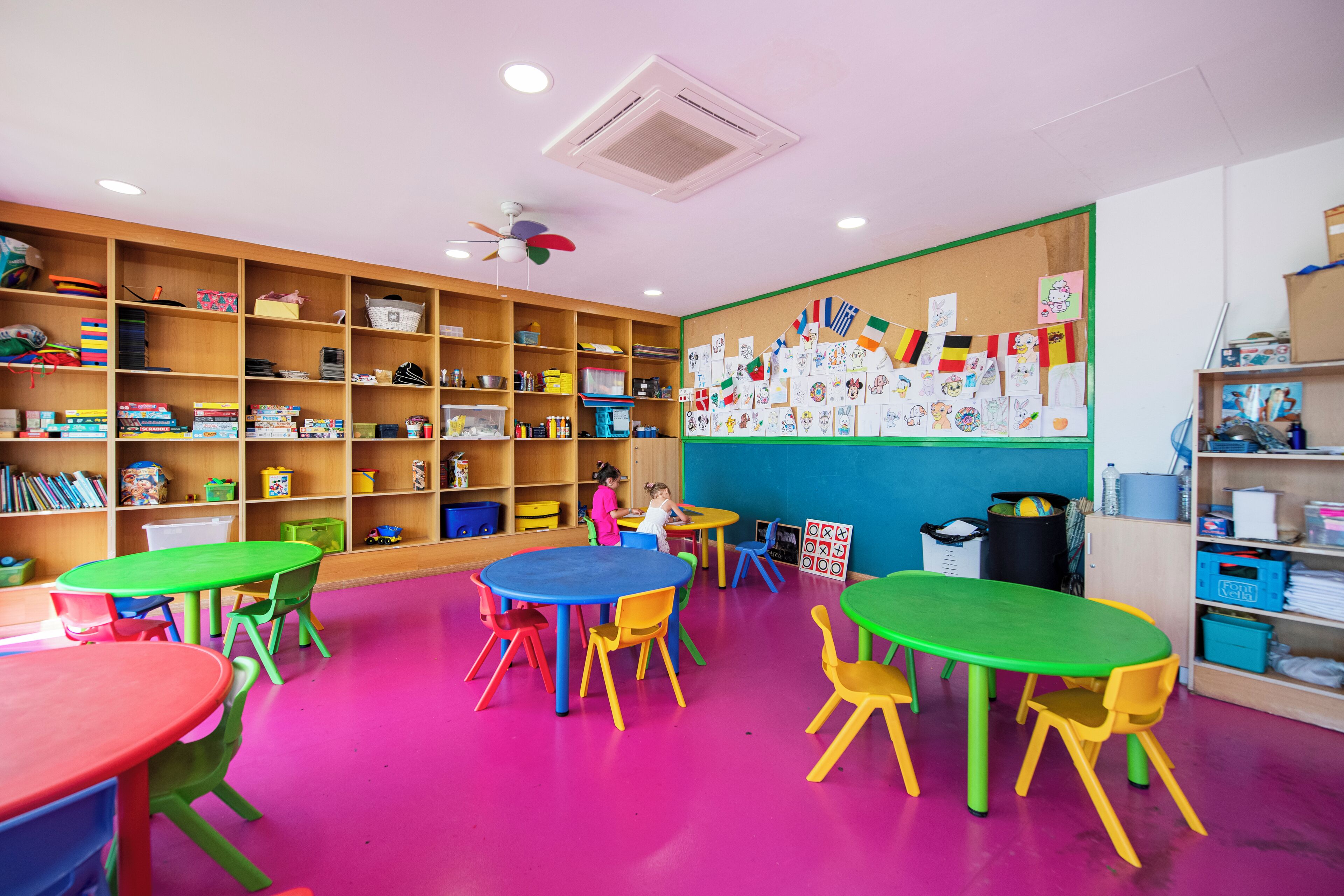 A classroom with colorful tables and chairs, shelves with educational materials, and a bulletin board with children's drawings.