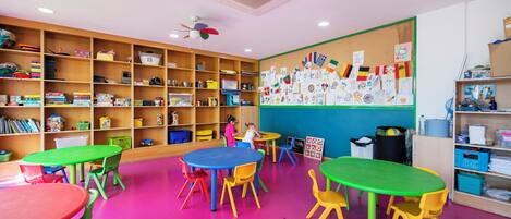 A classroom with colorful tables and chairs, shelves with educational materials, and a bulletin board with children's drawings.