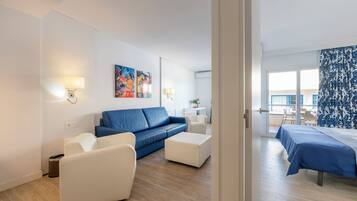 Una habitación de hotel moderna con un sofá azul, sillones blancos, una mesita y una cama con colcha azul.