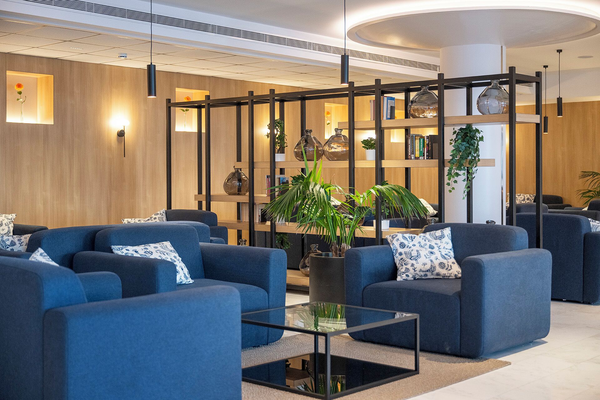 A modern lobby with sofas, a coffee table, and a bookshelf with plants.