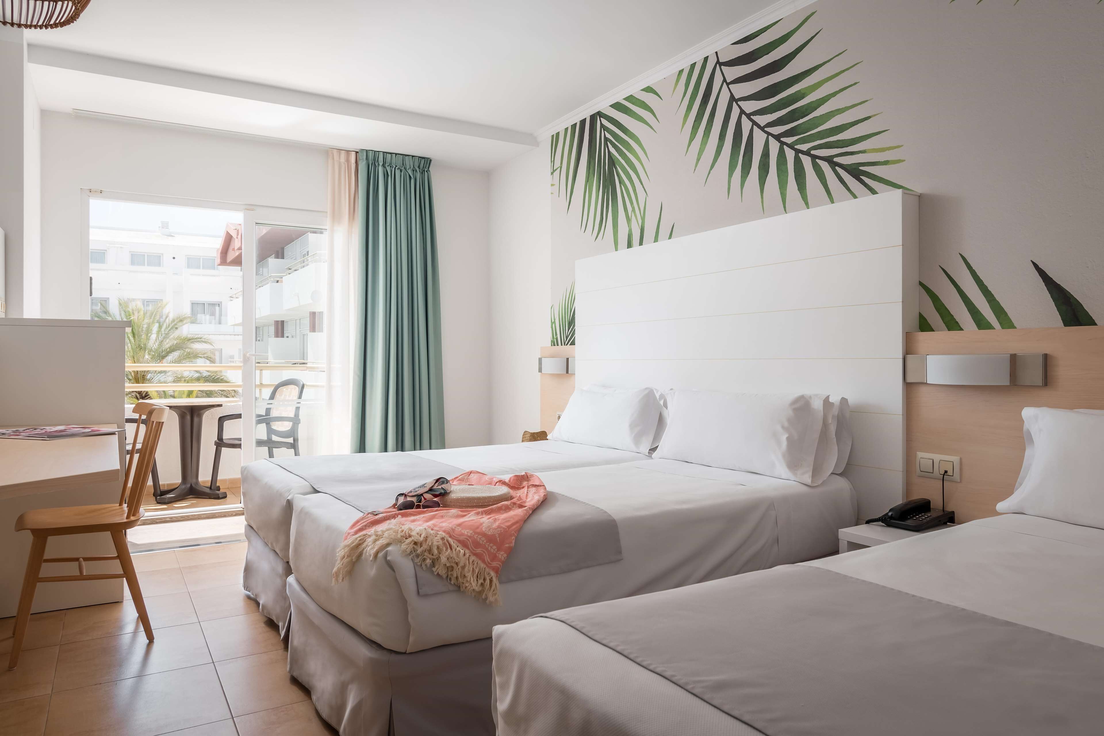 A hotel room with two beds, a desk, and a chair. There is a large window with a view of buildings outside.