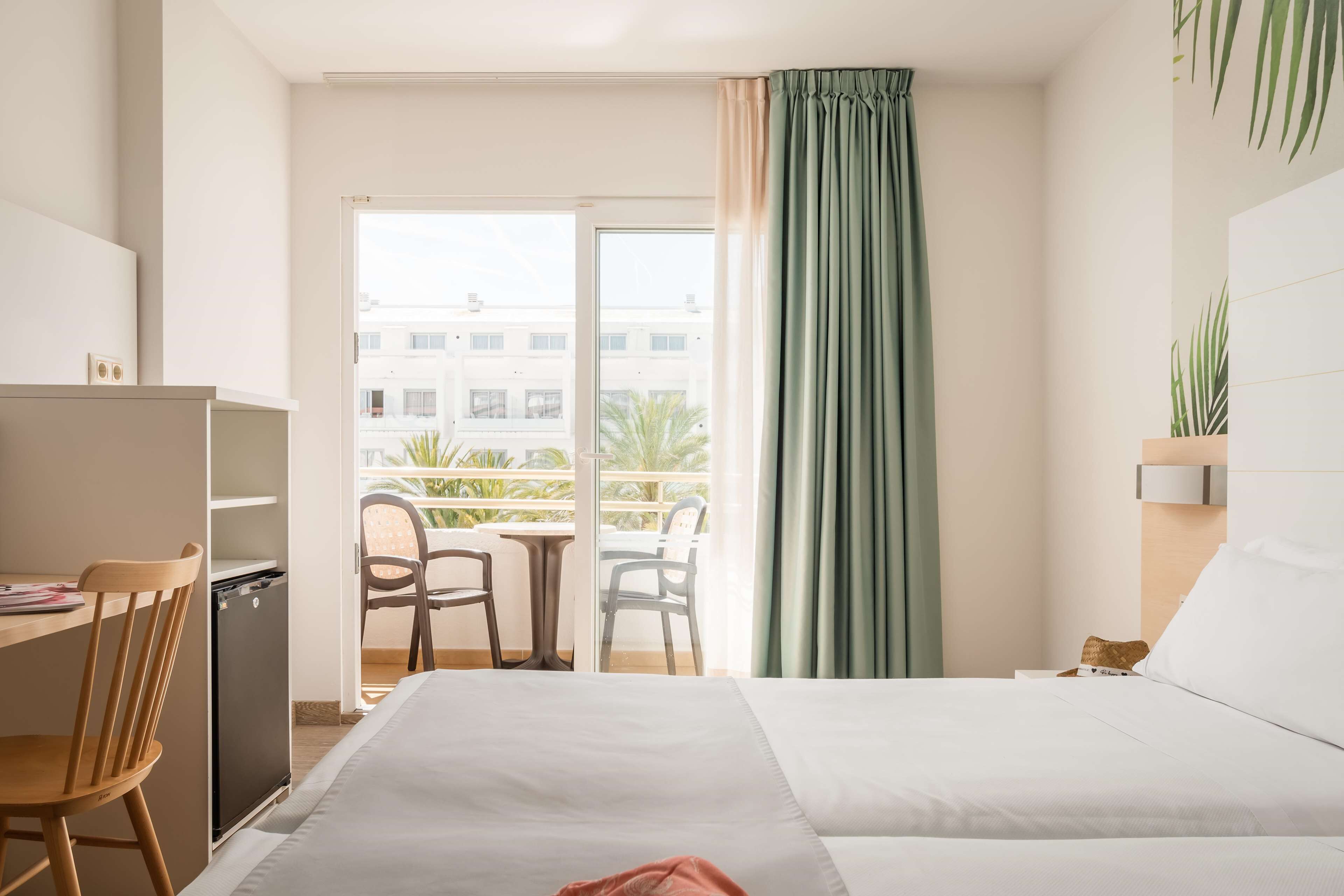 Une chambre d’hôtel comprenant un lit, un bureau avec une chaise, un petit réfrigérateur et un balcon avec une table et des chaises.