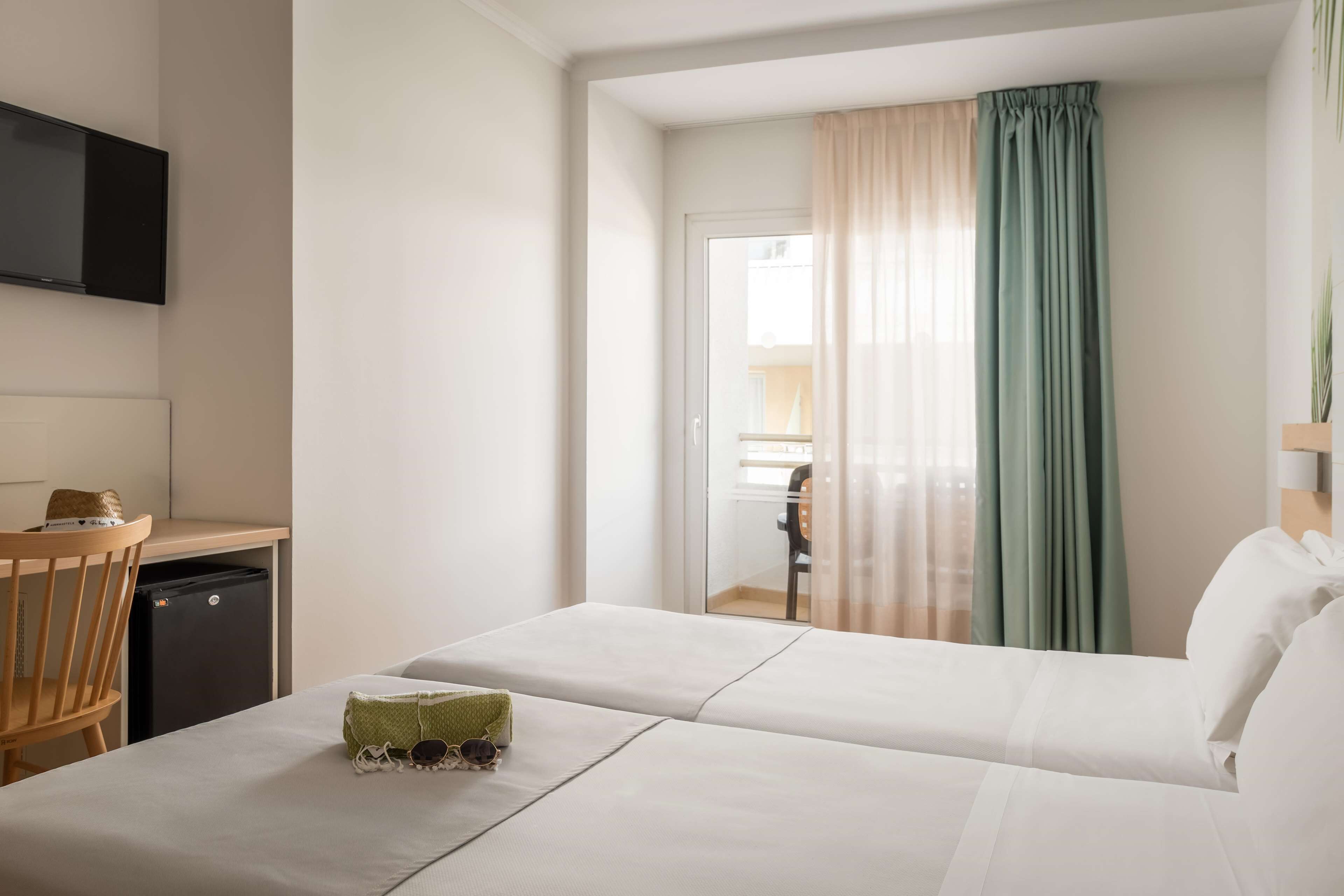 A hotel room with two beds, a desk, a television, and a view of the outside through a window with curtains.