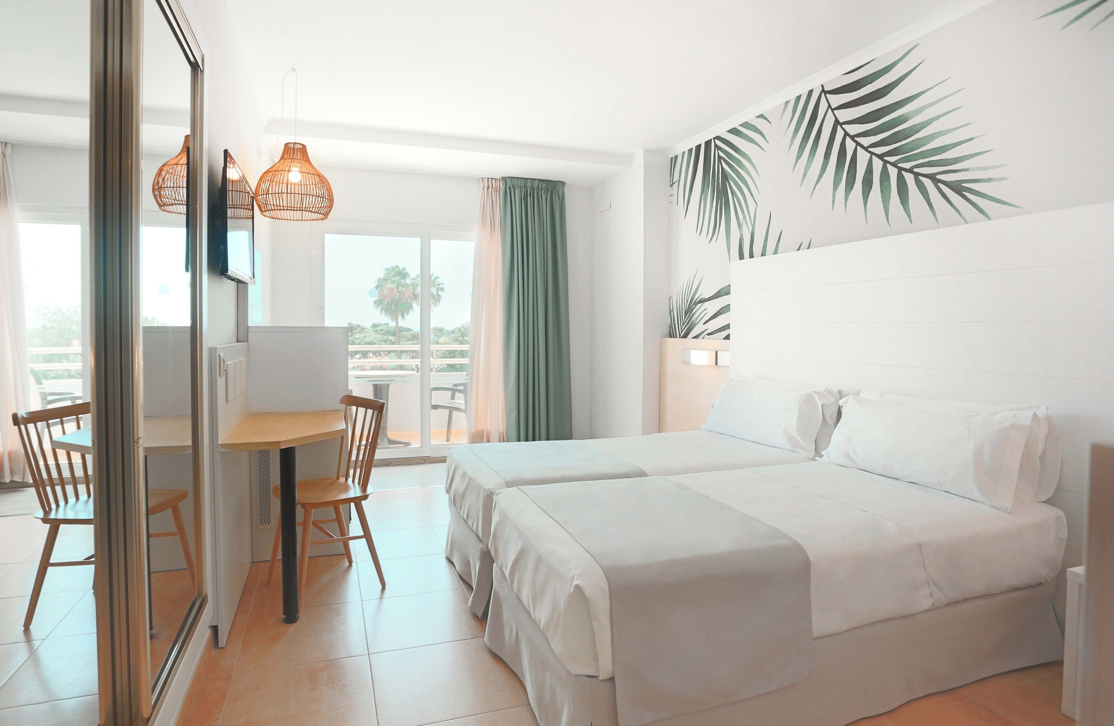 Hotelkamer met een bed, een bureau, stoelen, een raam met gordijnen en een wand met bladmotief.