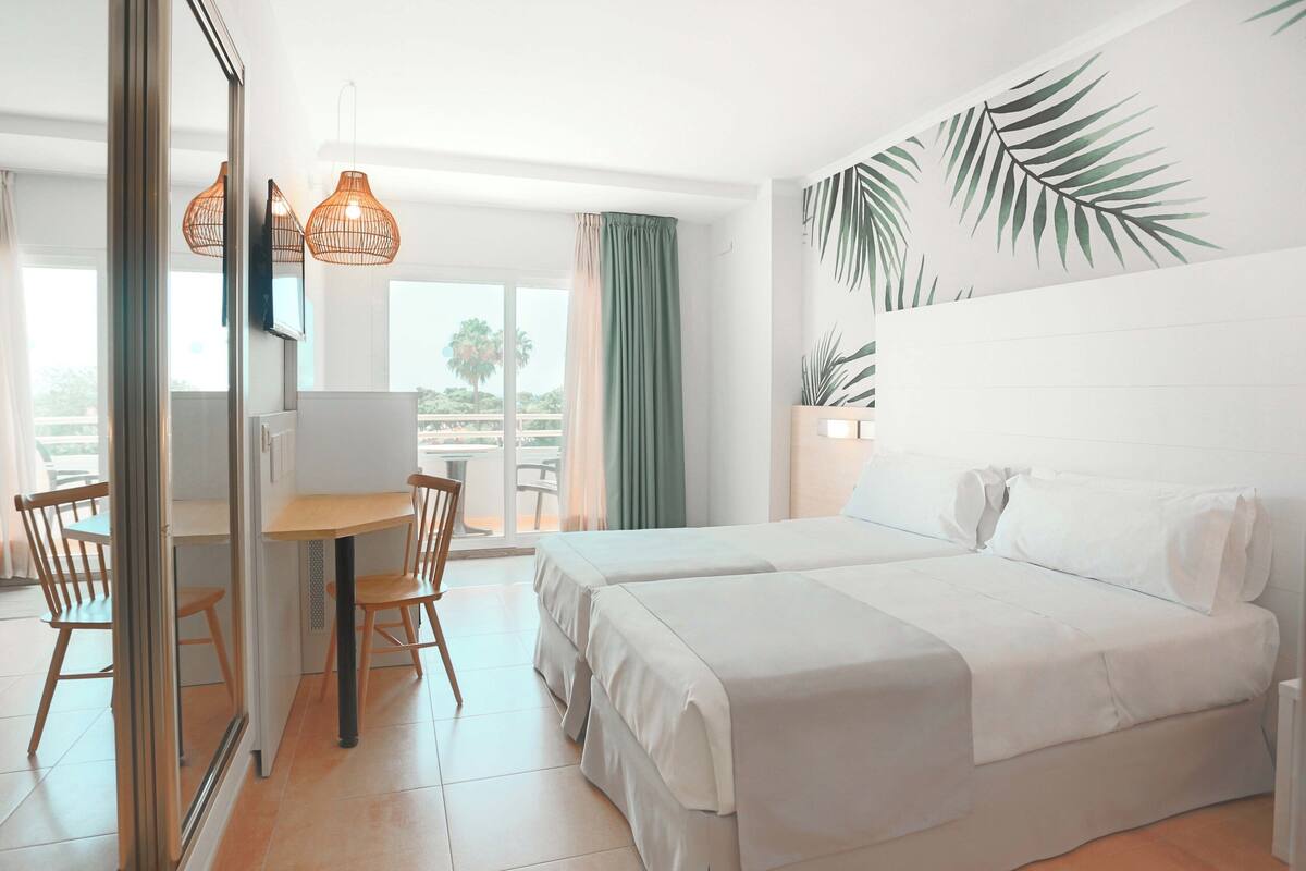 Une chambre d’hôtel comprenant un lit, un bureau, des chaises, une fenêtre avec des rideaux et un mur orné d’un motif de feuilles.