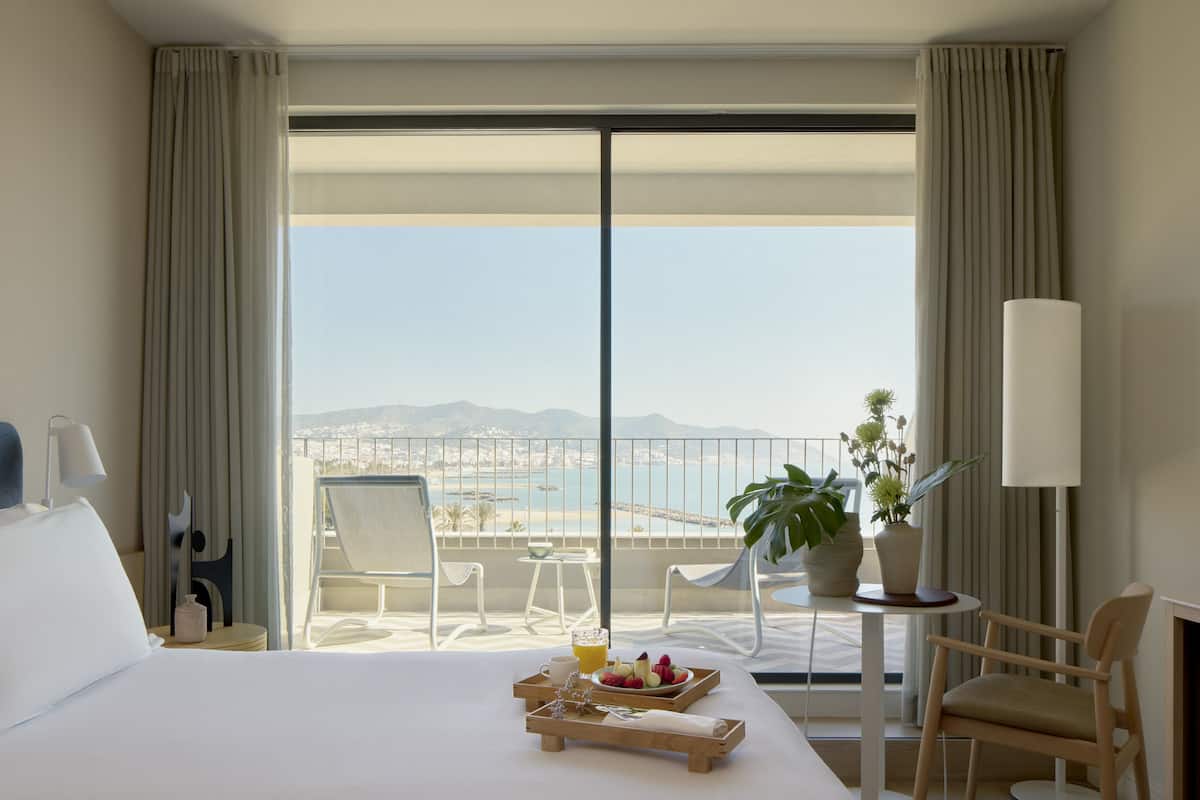 Ein Hotelzimmer mit Bett, Esstisch mit Obst und Tablett, Stuhl, Topfpflanze und Balkon mit Blick auf das Meer und die Berge.