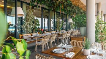 Un restaurante con mesas de madera y sillas de mimbre, rodeado de plantas en macetas y vegetación colgante.