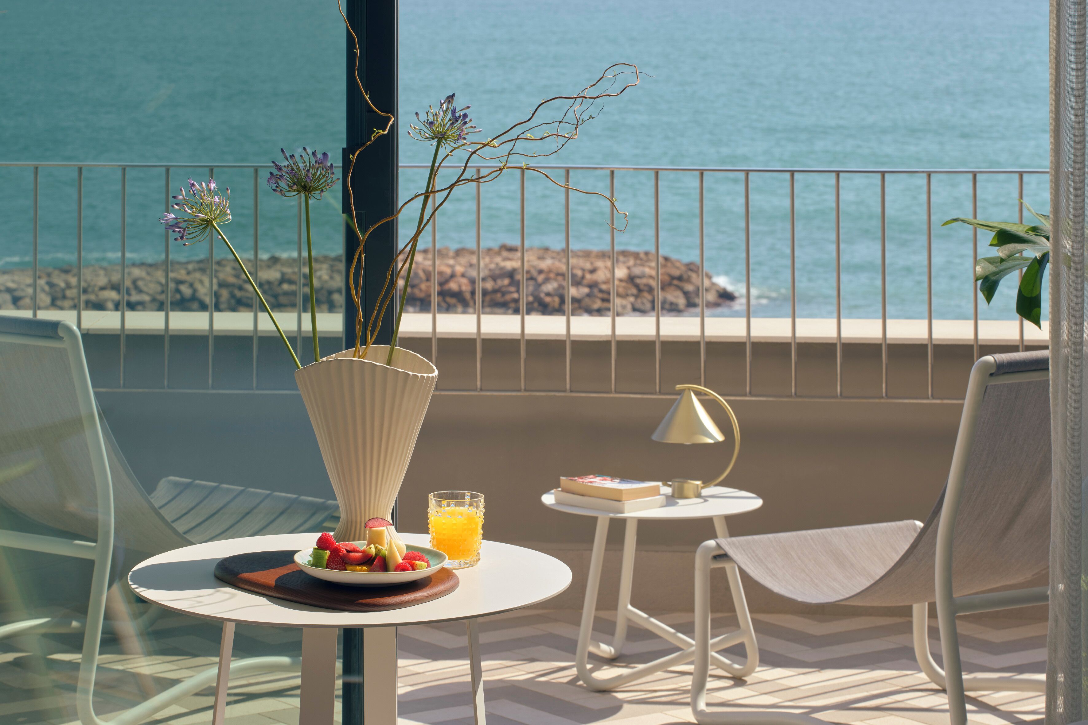 Un balcón con una mesa dispuesta para el desayuno, un jarrón con flores y vista al océano.