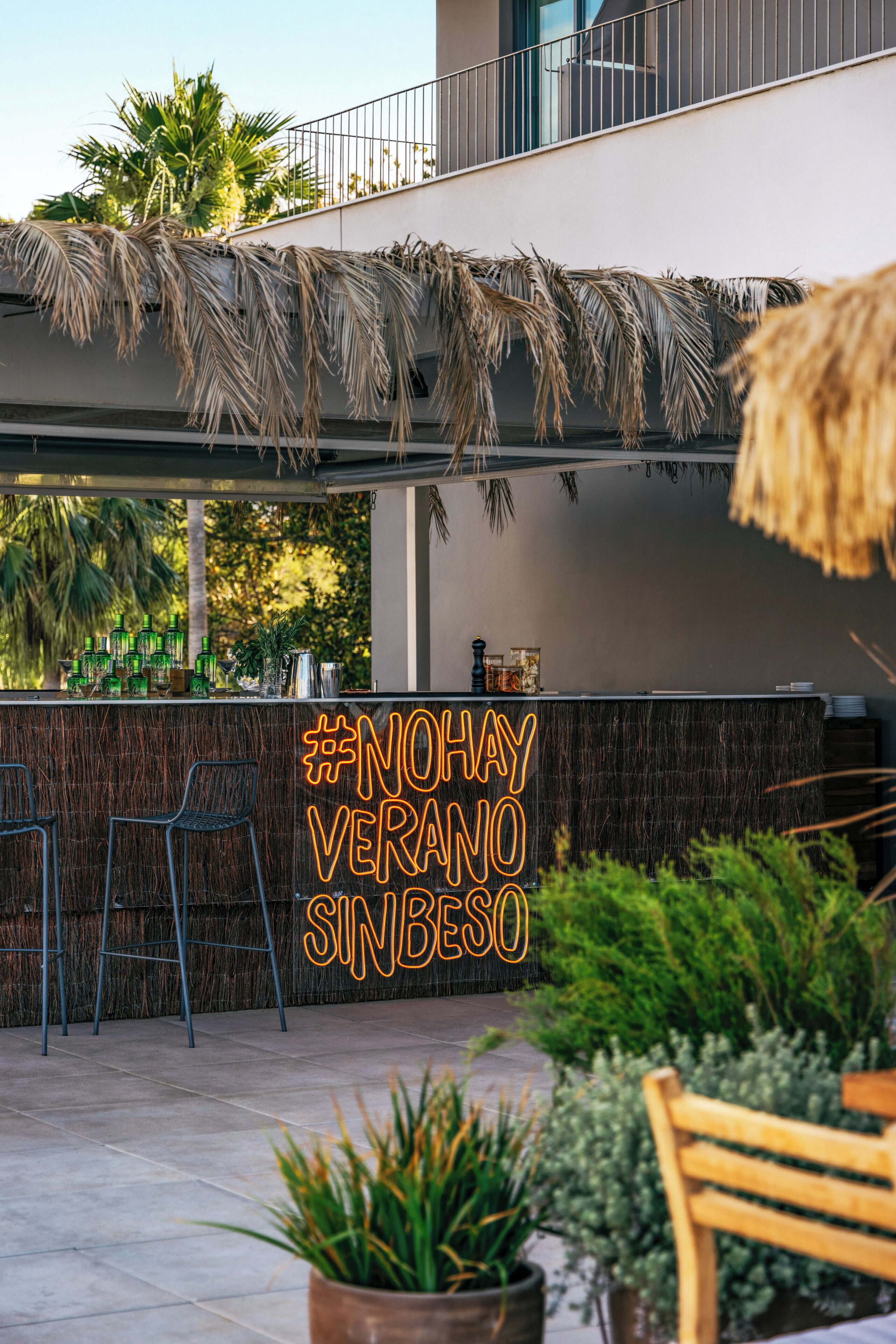 An outdoor bar area with a thatched roof, bar stools, and a neon sign reading "#NOHAYVERANO SINBESO".