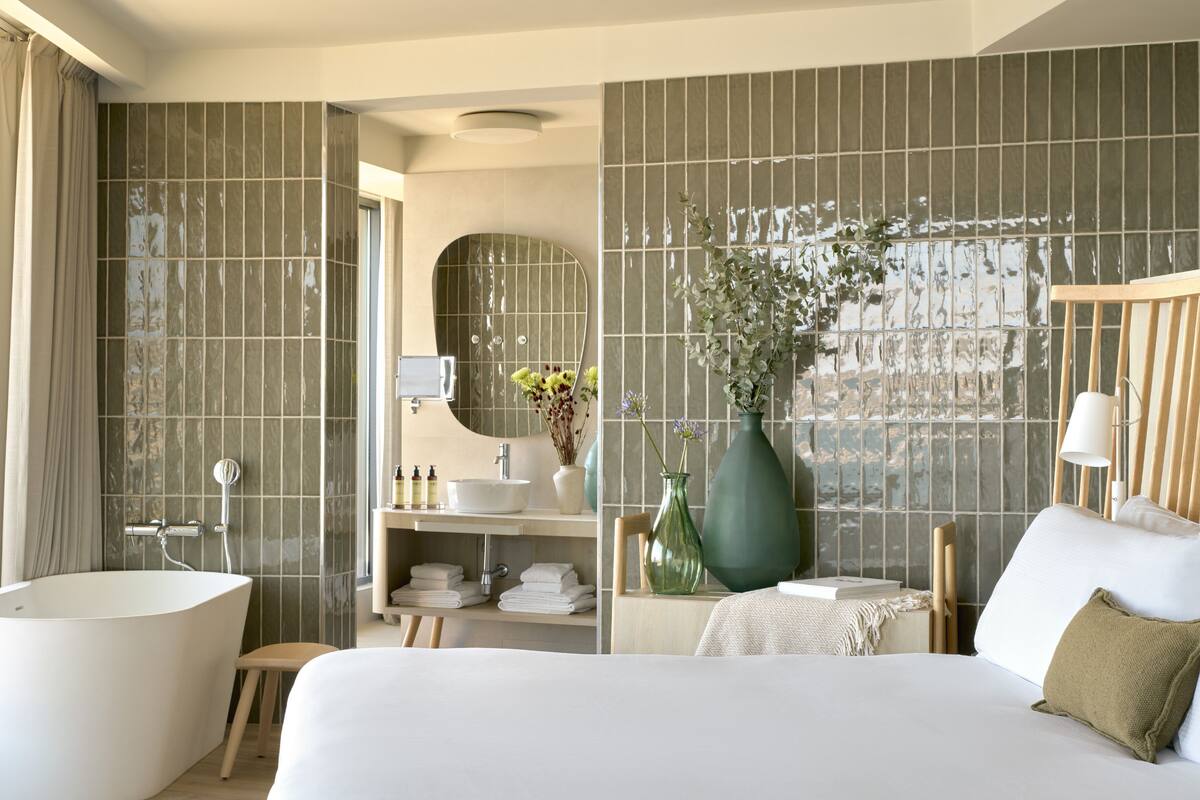 Ein modernes Hotelzimmer mit einem großen Bett, einer Badewanne, einem Schminktisch mit Spiegel und einer grünen Vase mit Blumen.