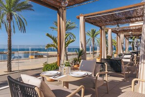 An outdoor dining area with tables set for two, overlooking a beach. - ME Sitges Terramar (Sitges)