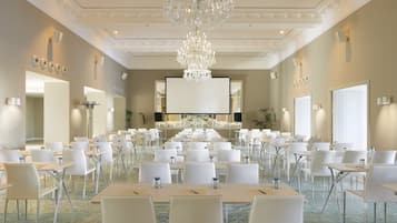 Un salón de conferencias amplio con filas de mesas y sillas blancas, una gran lámpara de araña y una pantalla grande al frente.