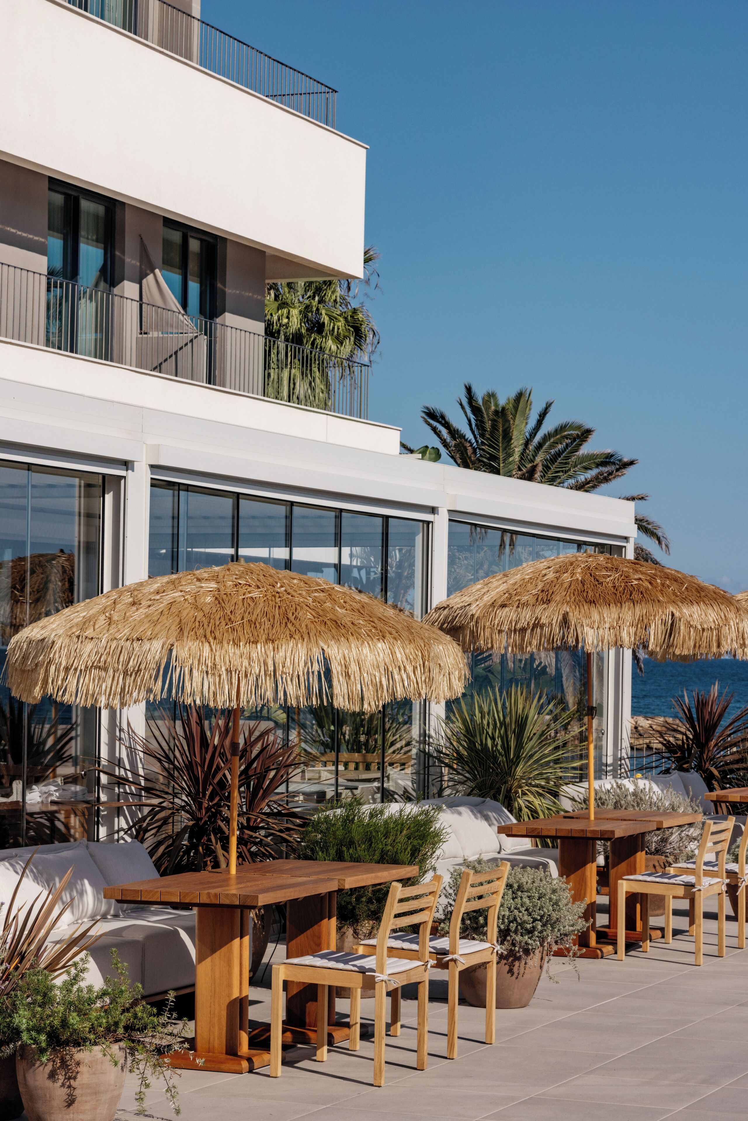 An outdoor seating area with wooden tables and chairs, thatched umbrellas, and potted plants.