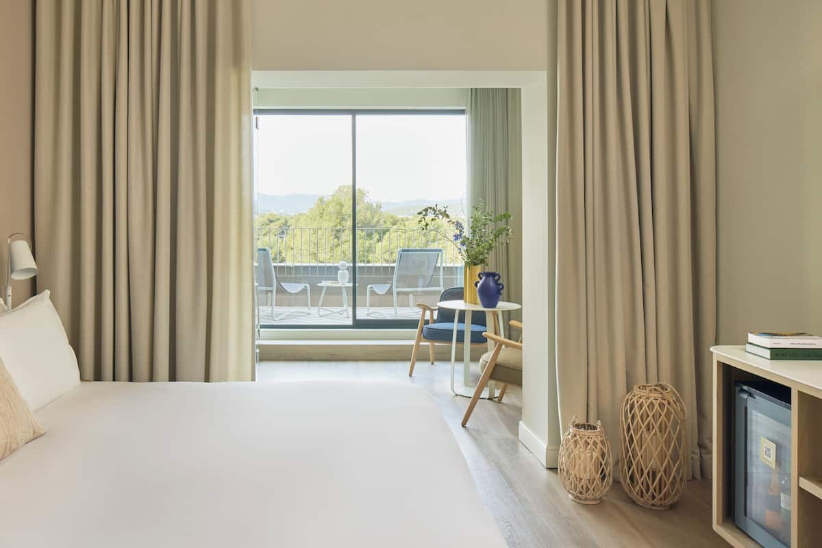 Ein Hotelzimmer mit einem großen Bett, einem Schreibtisch mit Stuhl, einer Topfpflanze und Blick ins Freie.