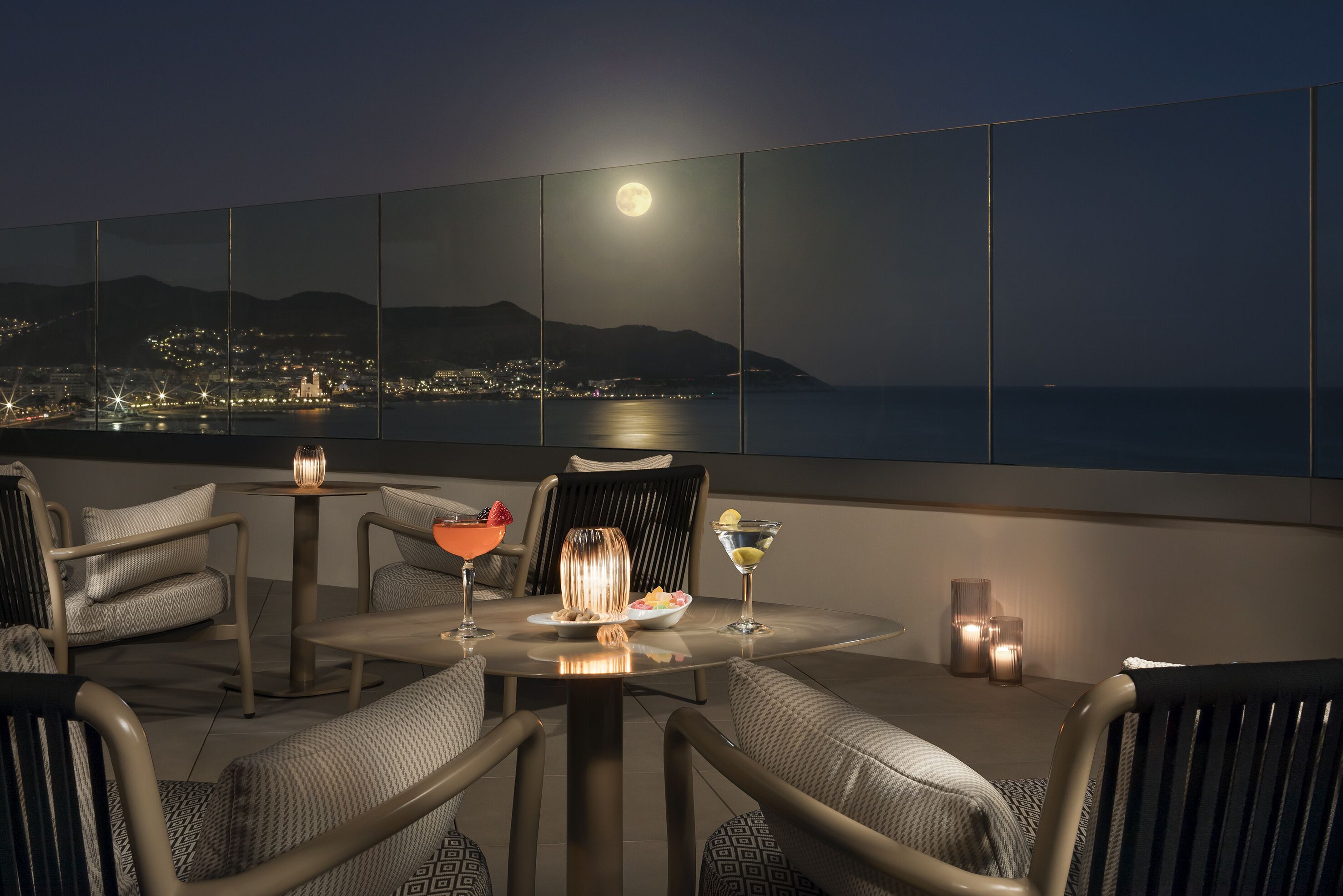 A rooftop terrace with a panoramic city view, a table with a fruit bowl, a cocktail, and candles.