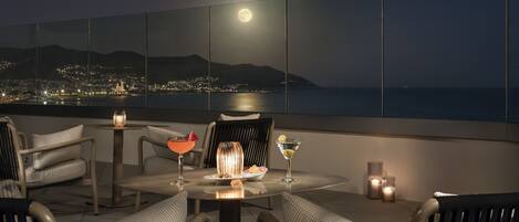 A rooftop terrace with a panoramic city view, a table with a fruit bowl, a cocktail, and candles.