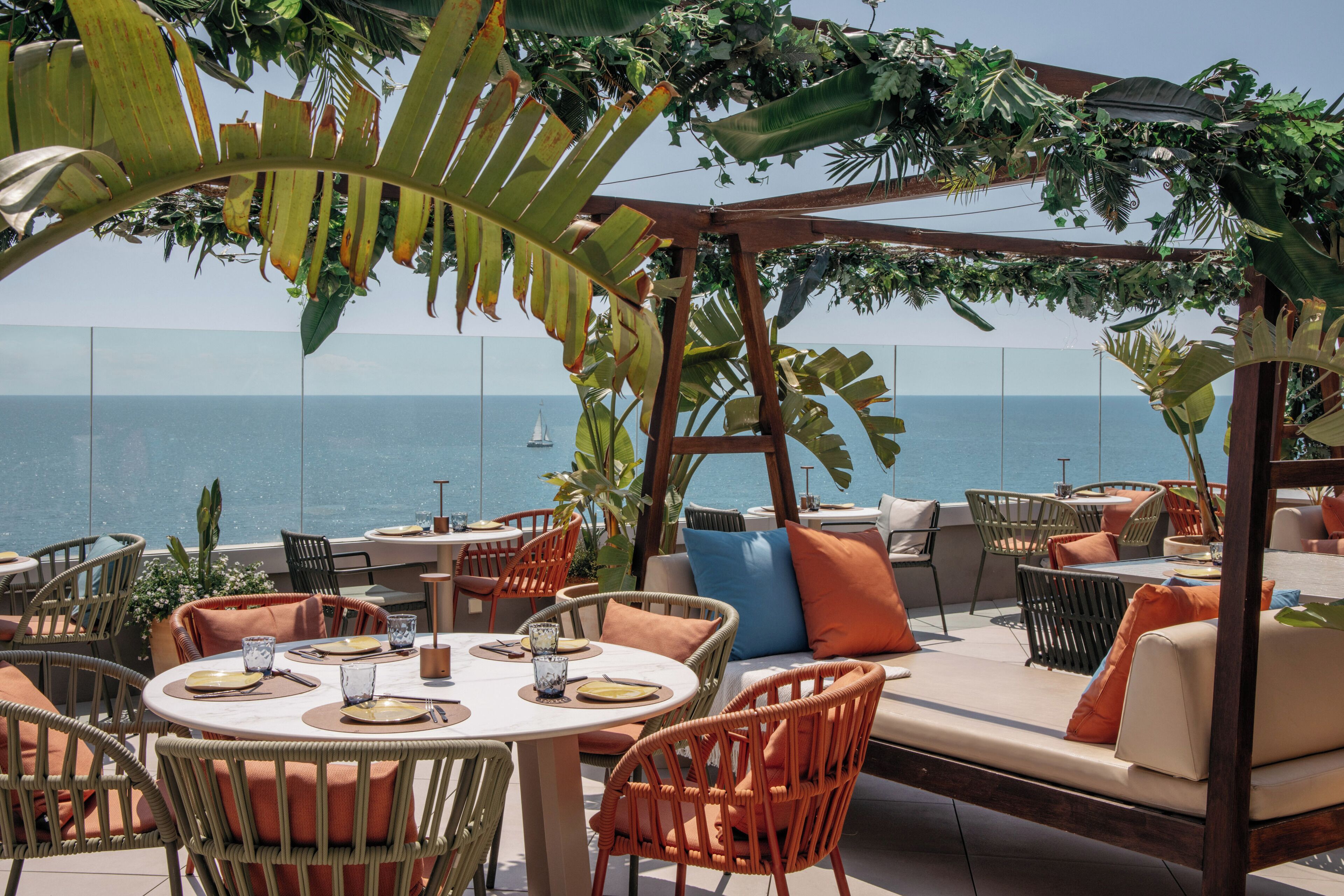 Un área de comedor al aire libre con mobiliario de mimbre, vistas al mar y una pérgola con plantas trepadoras.