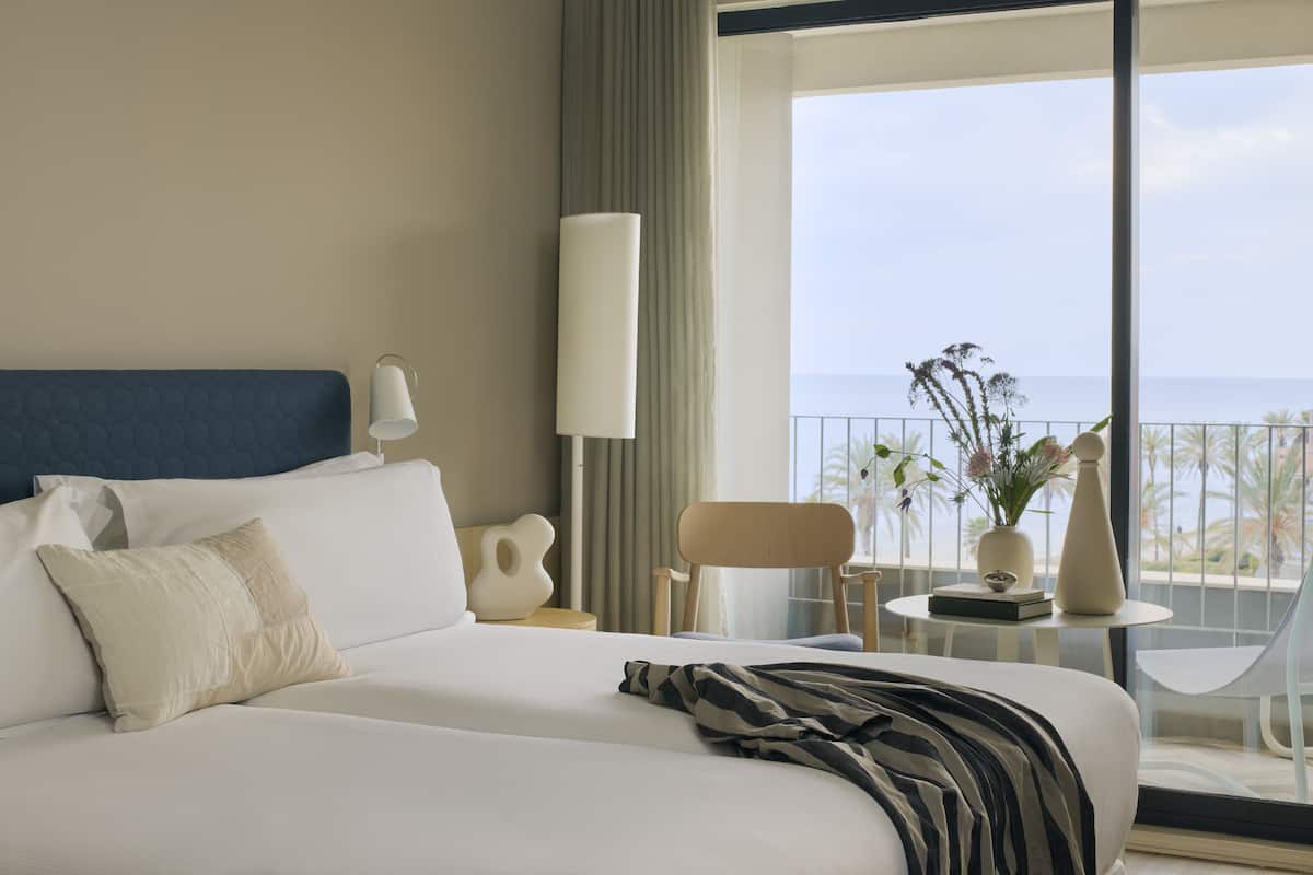 Ein Hotelzimmer mit einem großen Bett, einem Nachttisch mit Lampe, einem Sessel und Blick aufs Meer.