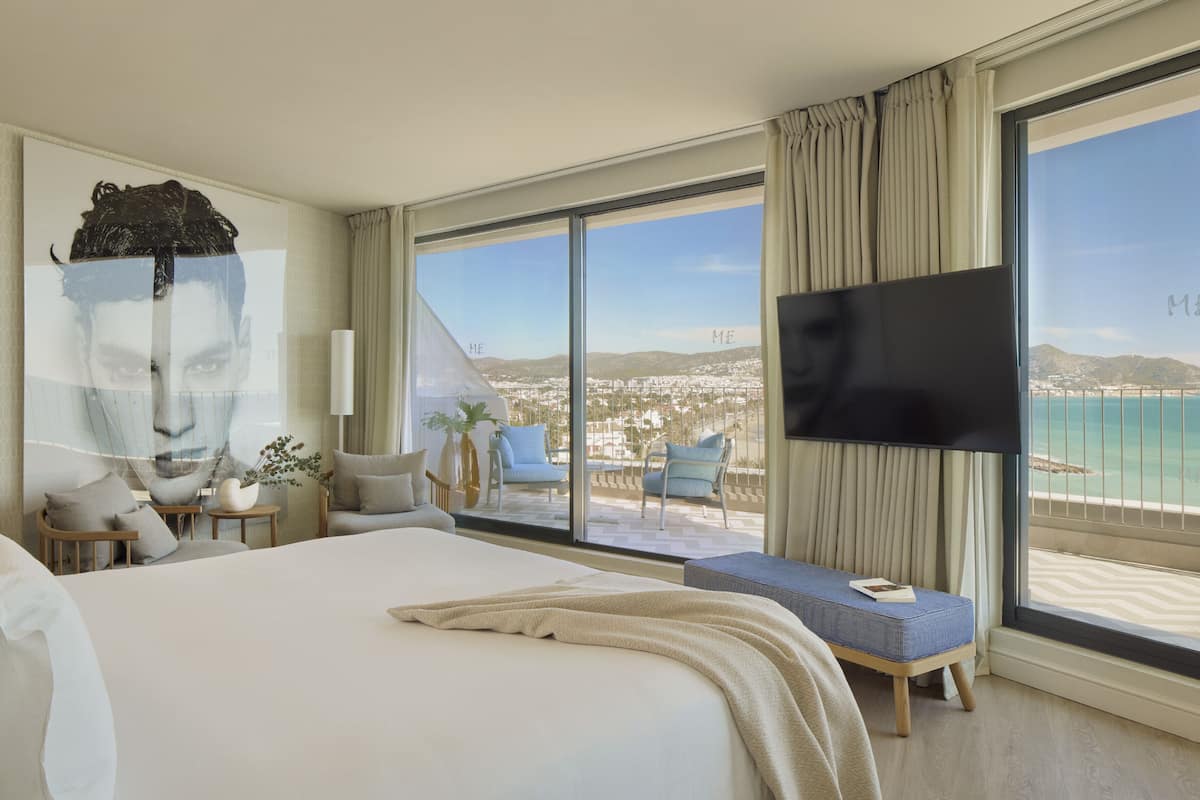 Ein Hotelzimmer mit einem großen Bett, einem Fernseher, einem Balkon mit Meerblick und einem großen Fenster.