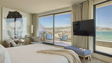 A hotel room with a large bed, a flat-screen TV, a balcony with a view of the sea, and a large window.