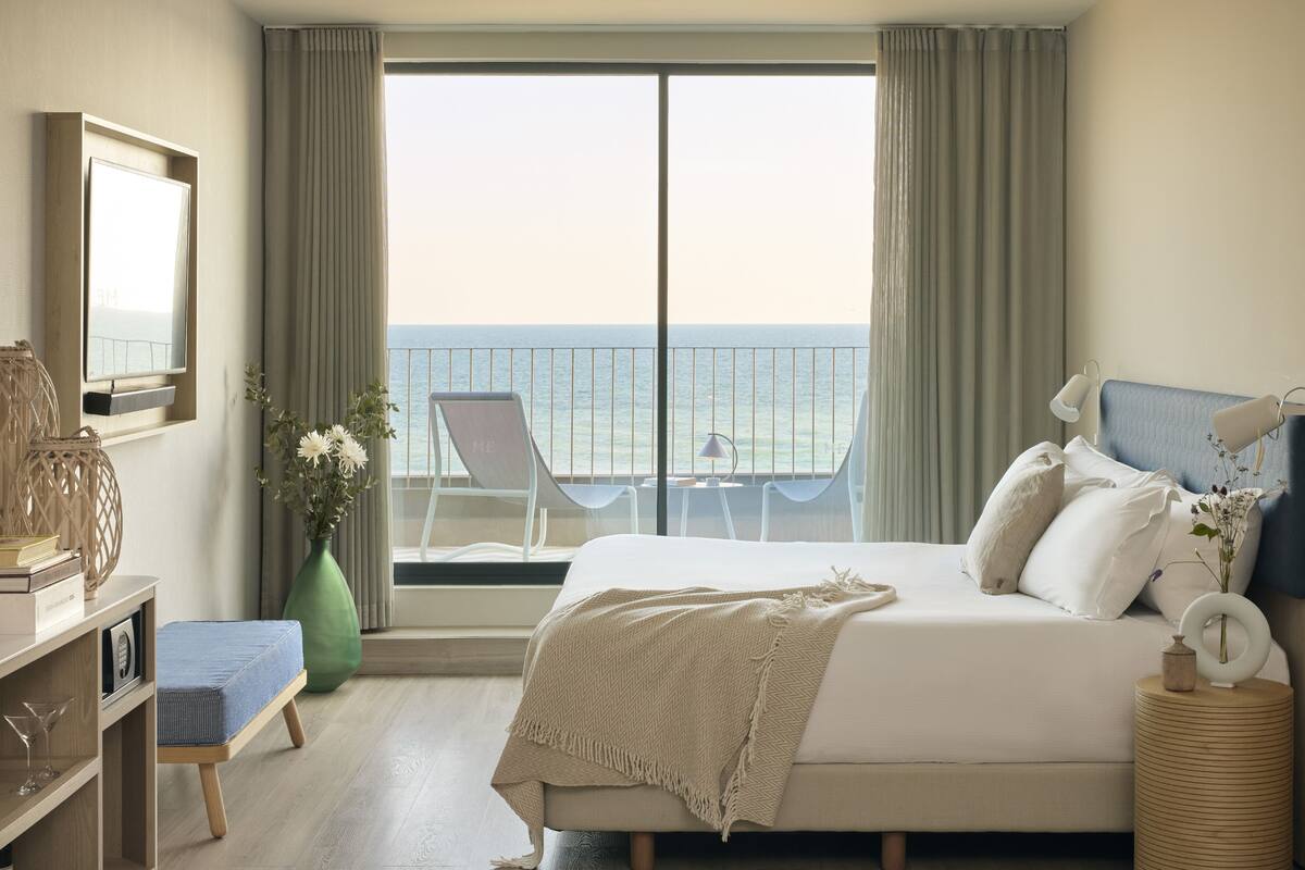 Ein Schlafzimmer mit einem großen Bett, einem Schreibtisch, einem Stuhl, einer Vase mit Blumen und Meerblick vom Balkon.