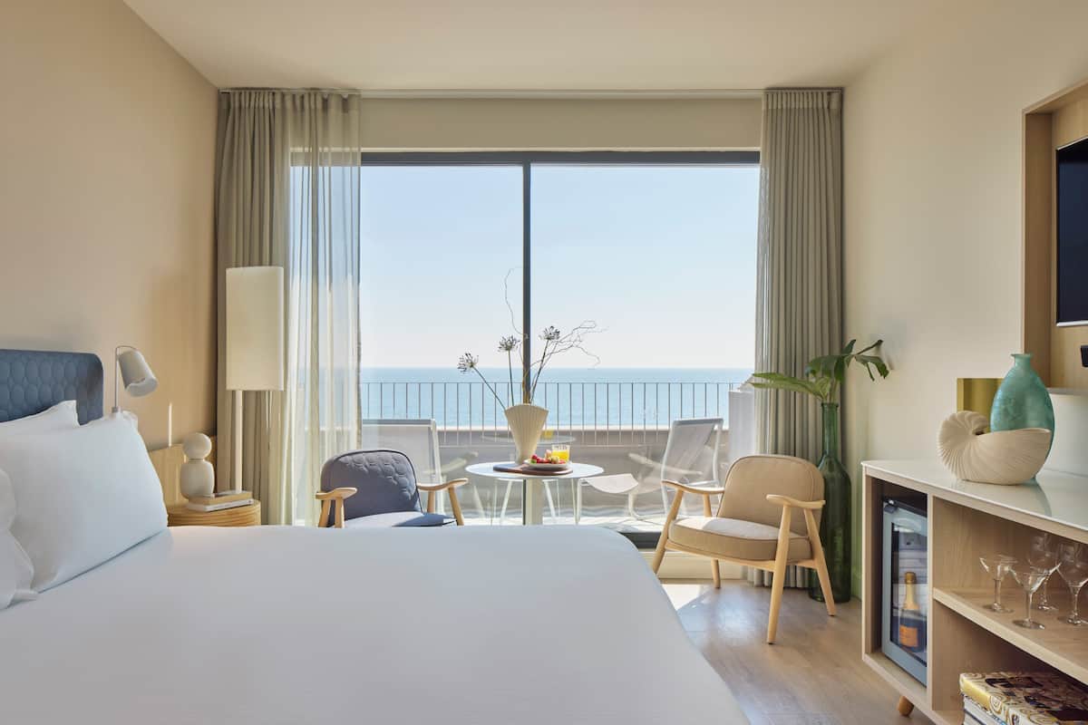 Ein Hotelzimmer mit einem großen Bett, einem Balkon mit Blick, einem kleinen Tisch mit einer Vase und einem Fernseher an der Wand.