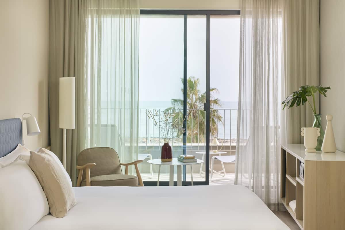 Ein Hotelzimmer mit einem großen Bett, einem Sessel, einem kleinen Tisch mit einer Vase und Blick auf das Meer durch transparente Vorhänge.