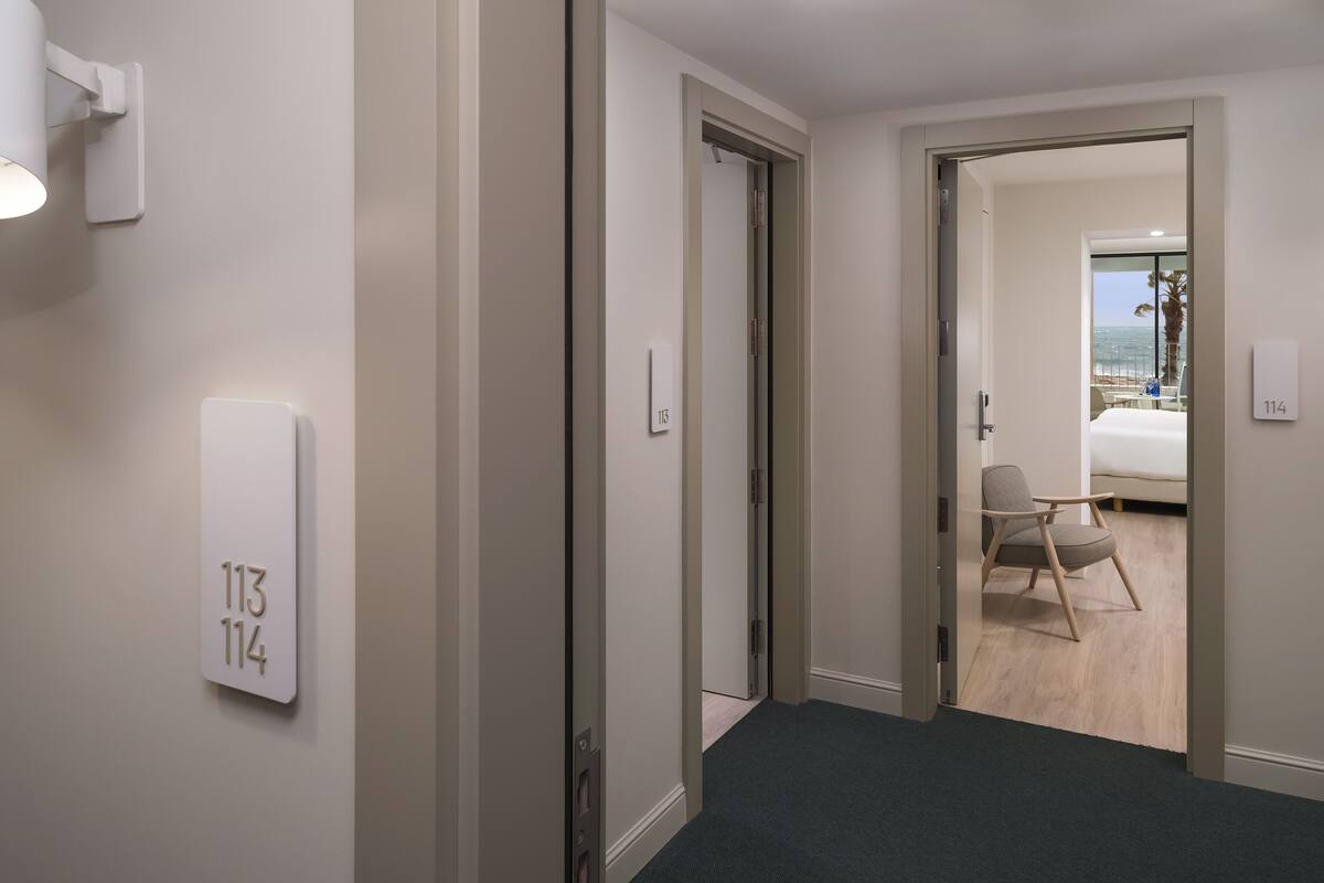 Ein Hotelgang mit einer Tür zu einem Zimmer, das ein Bett, einen Stuhl und Meerblick bietet.
