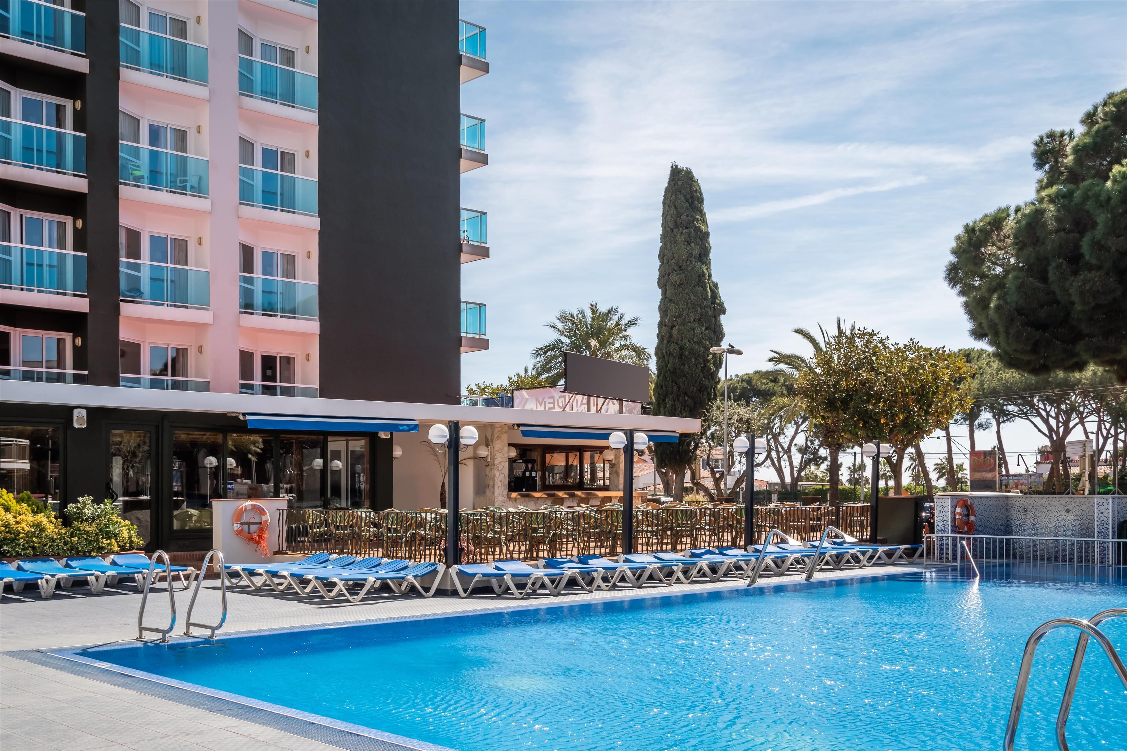 Una piscina con sillones de descanso y una zona de bar, rodeada por un edificio de varios pisos y balcones.