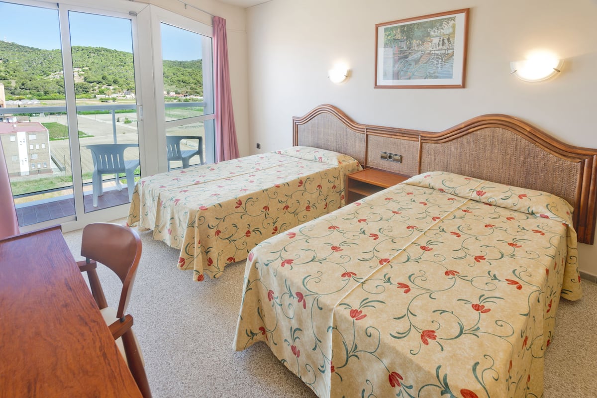 Ett hotellrum med två sängar, ett skrivbord i trä, en stol och en balkong med utsikt över ett grönskande landskap.