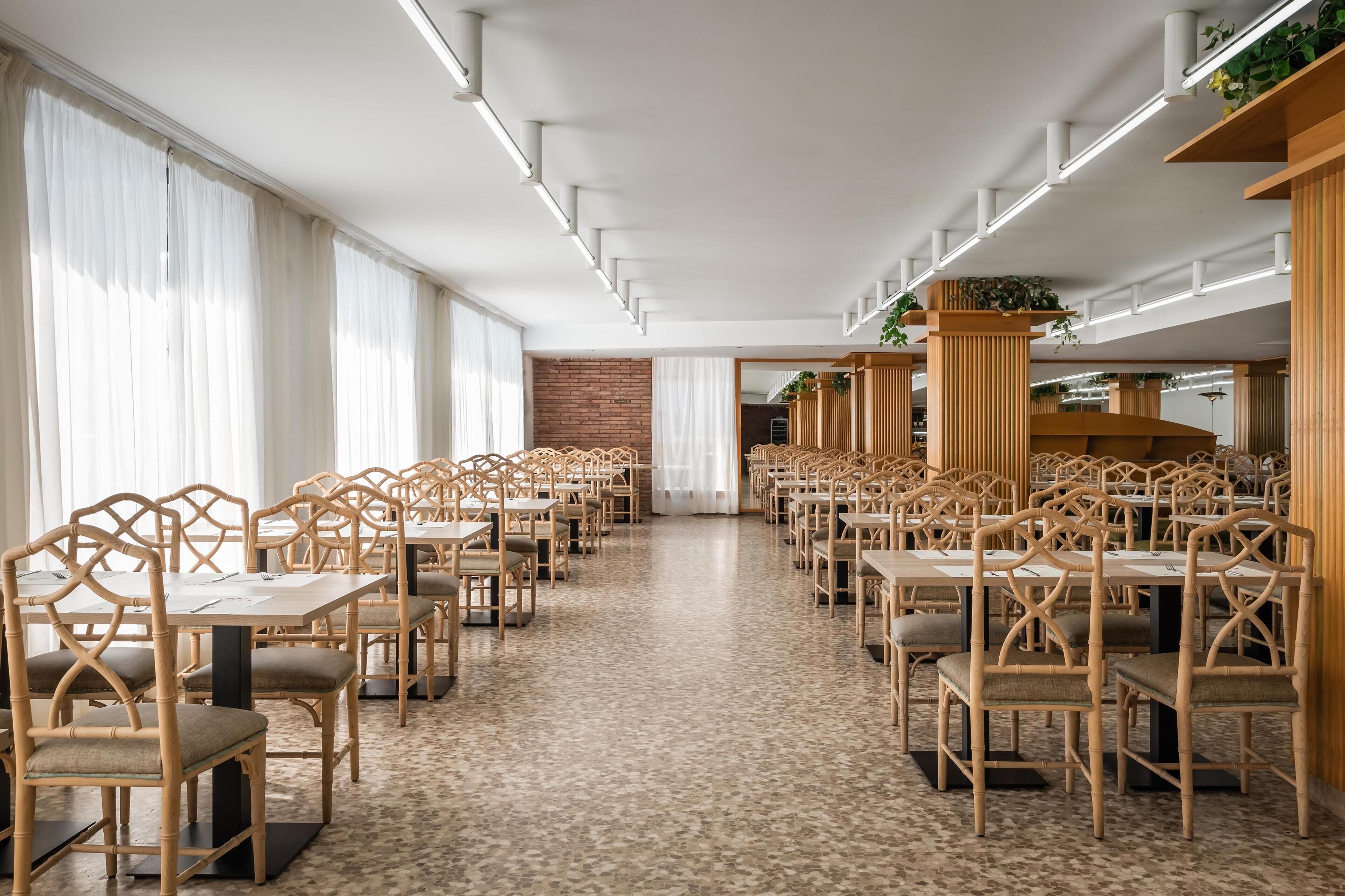 A spacious dining area with long tables and chairs, large windows with sheer curtains, and a brick wall in the background.