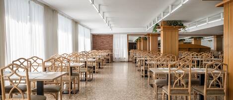 A spacious dining area with long tables and chairs, large windows with sheer curtains, and a brick wall in the background.