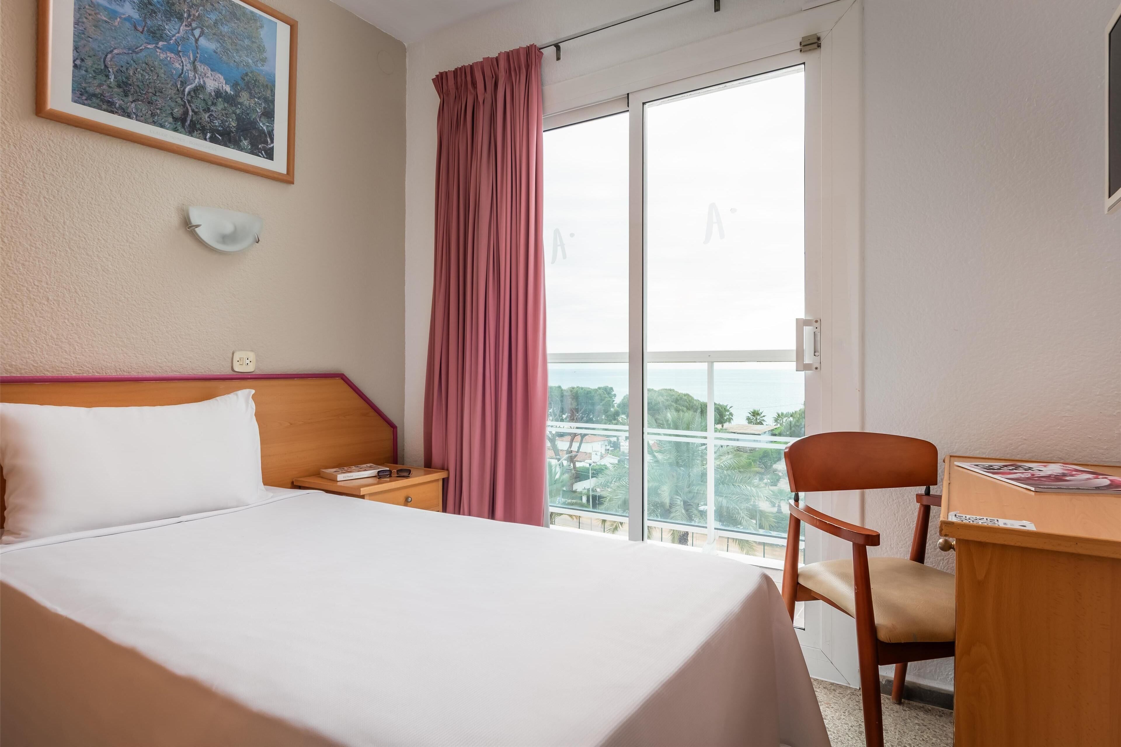 A hotel room with a bed, a desk, a chair, a window with a view, and a painting on the wall.