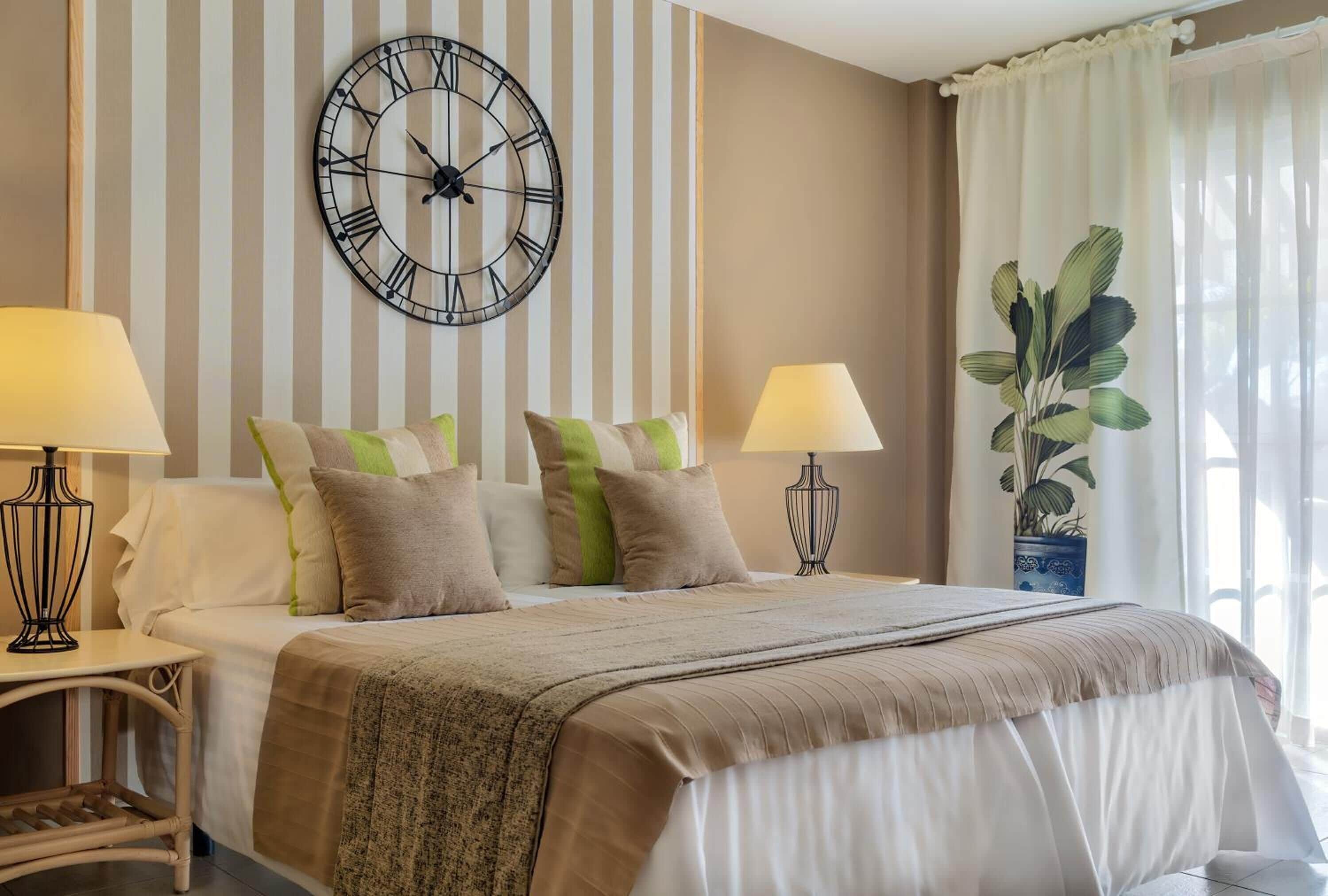 Una habitación de hotel con una cama grande, dos lámparas de noche, un reloj de pared y una planta en maceta.
