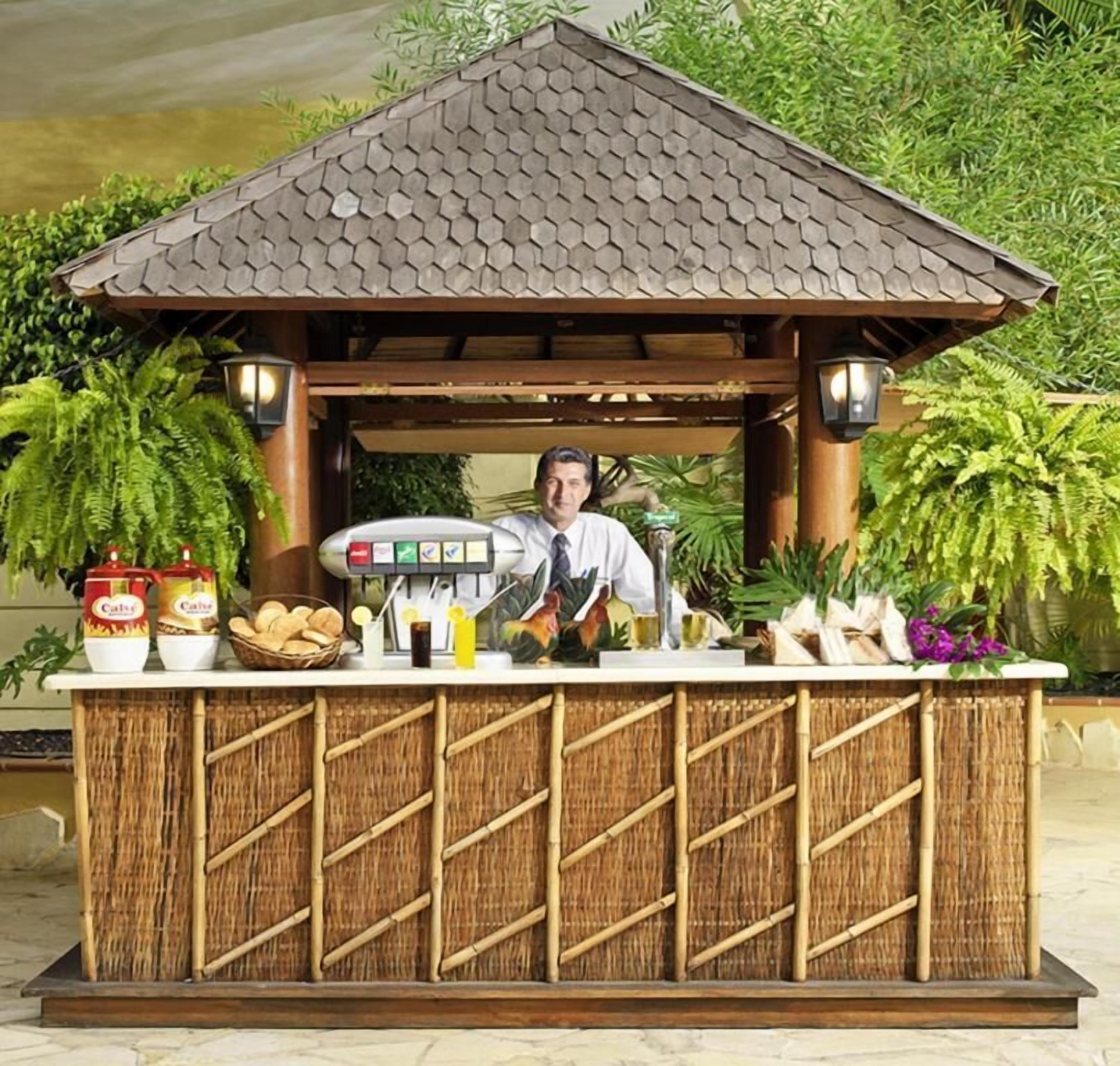 Un bar tiki con techo de paja, una barra con diversas bebidas y aperitivos, y un fondo de exuberante follaje.