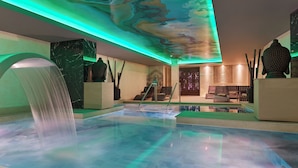 Un espace piscine intérieur moderne, agrémenté d’une cascade, de sièges et d’un plafond décoratif.