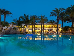 A resort pool area with lounge chairs, palm trees, and a building with multiple levels and outdoor seating. - Gran Oasis Resort (Arona)