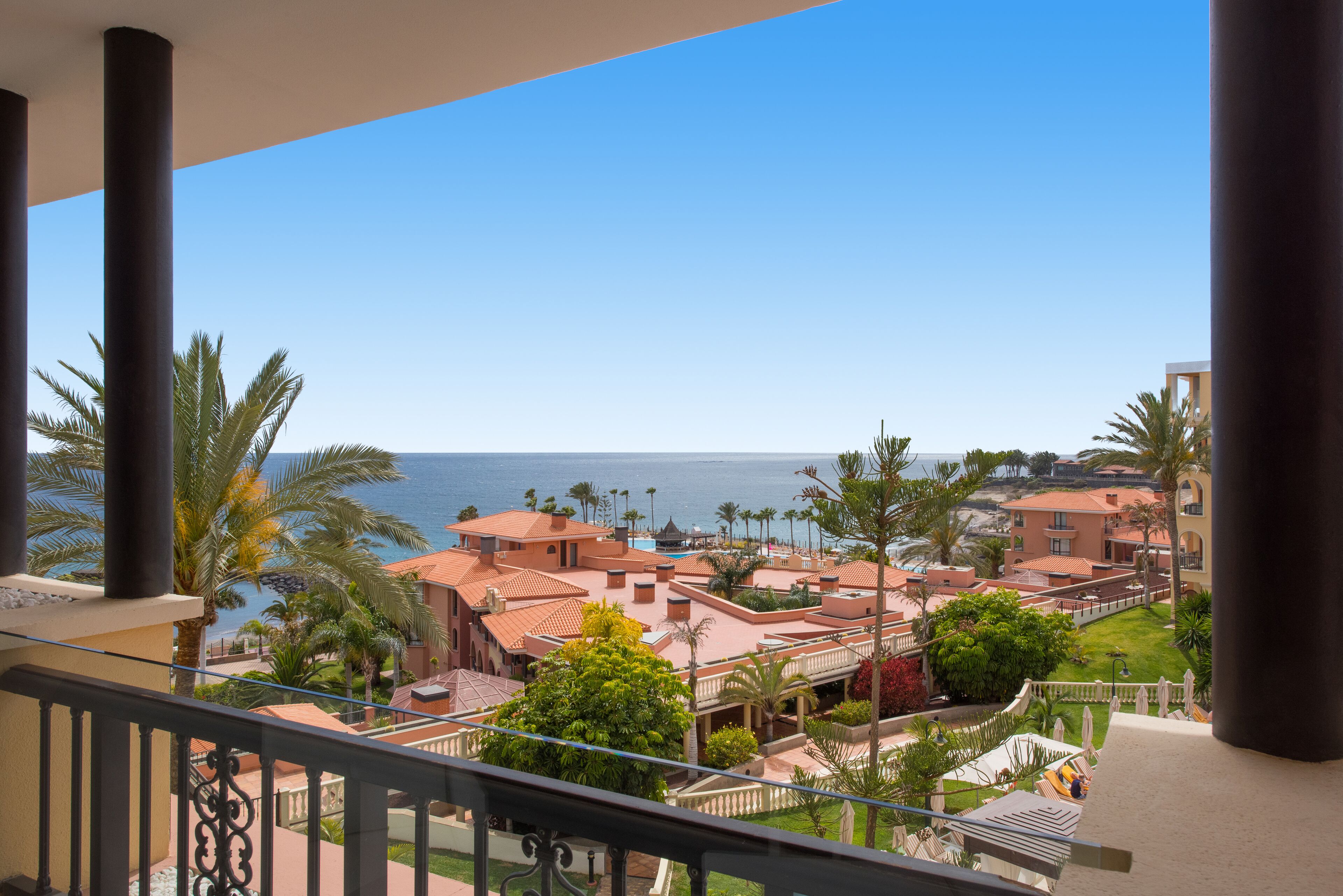 A balcony with a view of a coastal resort area, featuring palm trees, a pool, and buildings with red-tiled roofs.