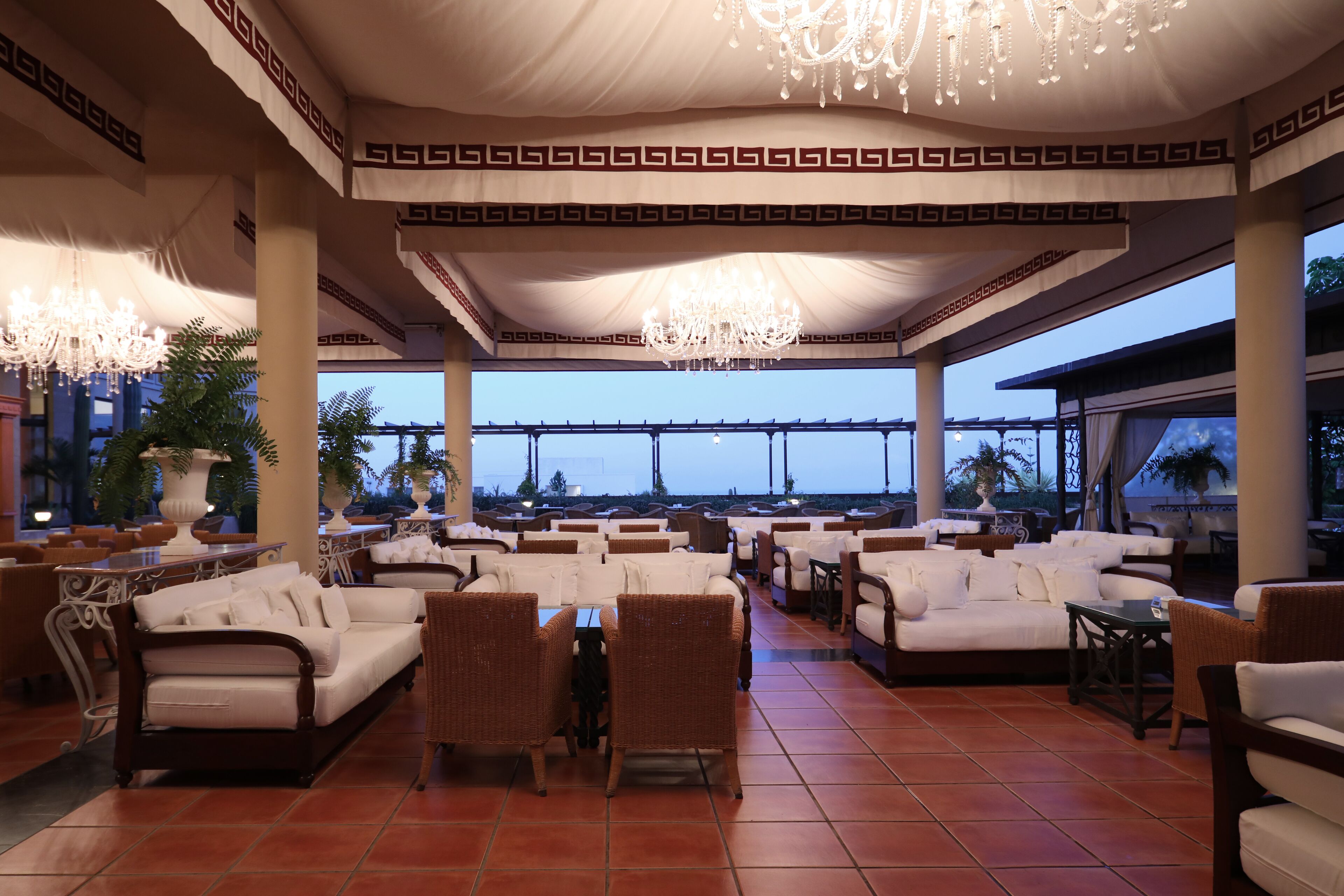 A spacious outdoor seating area with white sofas and wicker chairs, a tiled floor, and a view of the ocean.