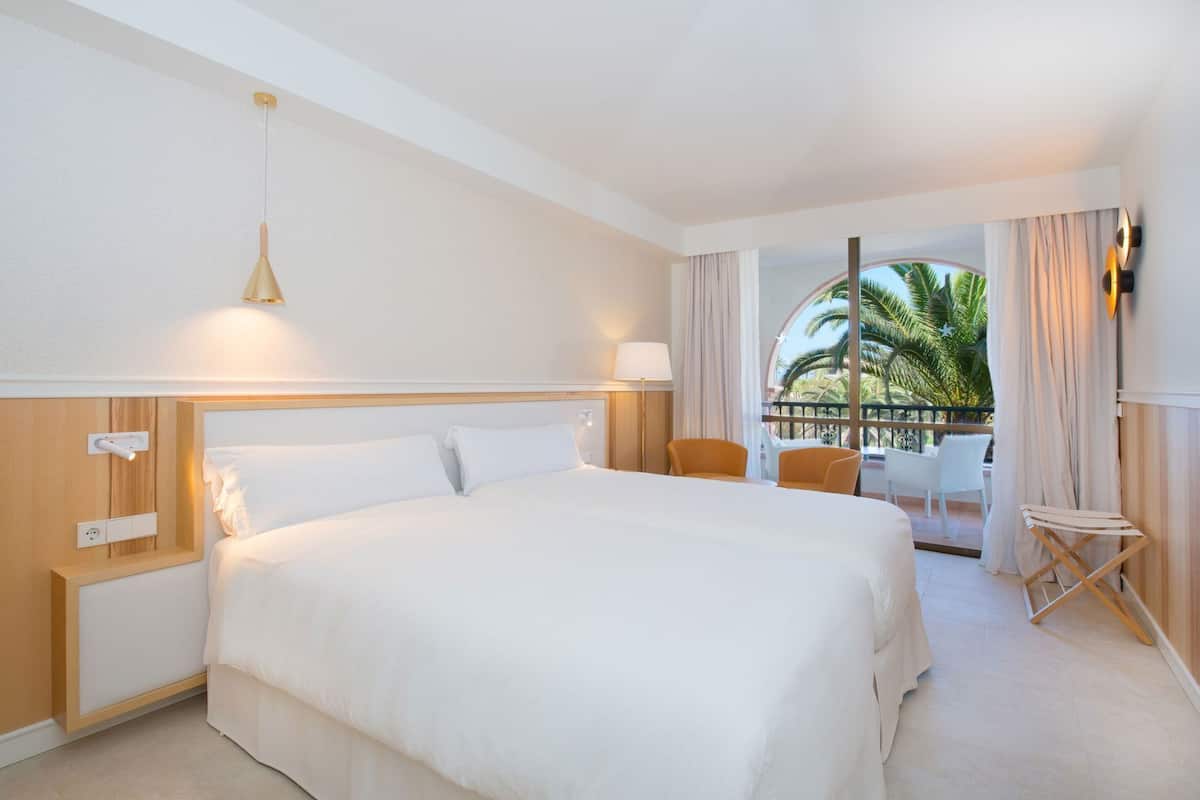 Quarto de hotel com uma cama grande, uma mesa pequena, duas cadeiras, uma varanda com vista para palmeiras e um lustre.