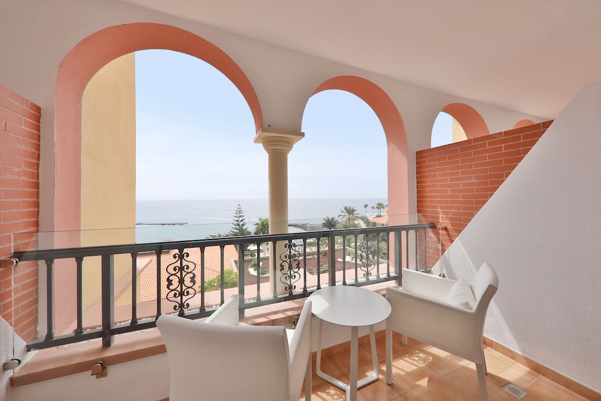 A balcony with white chairs and a table, overlooking a coastal view with a clear sky.