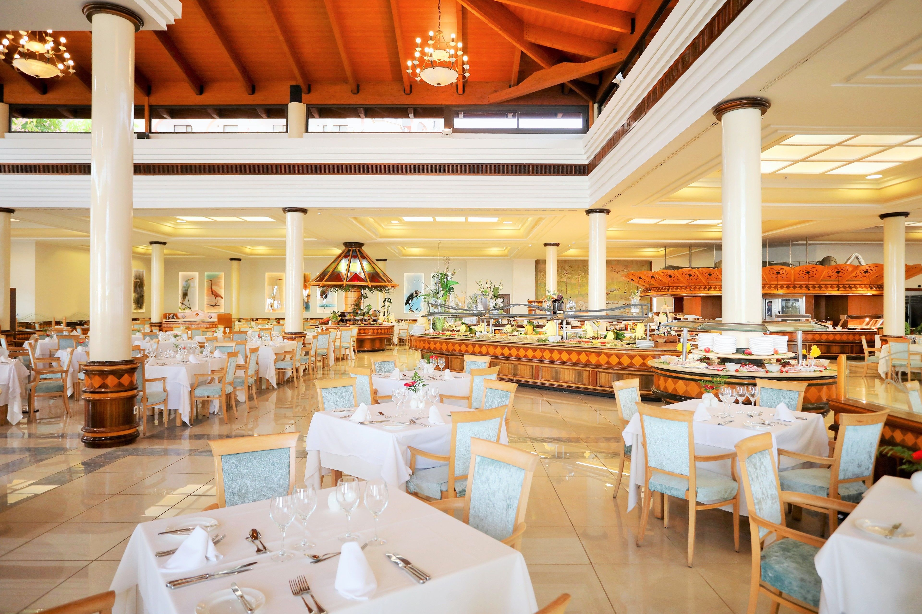 A spacious dining area with multiple tables and chairs, a central buffet, and a high ceiling with chandeliers.